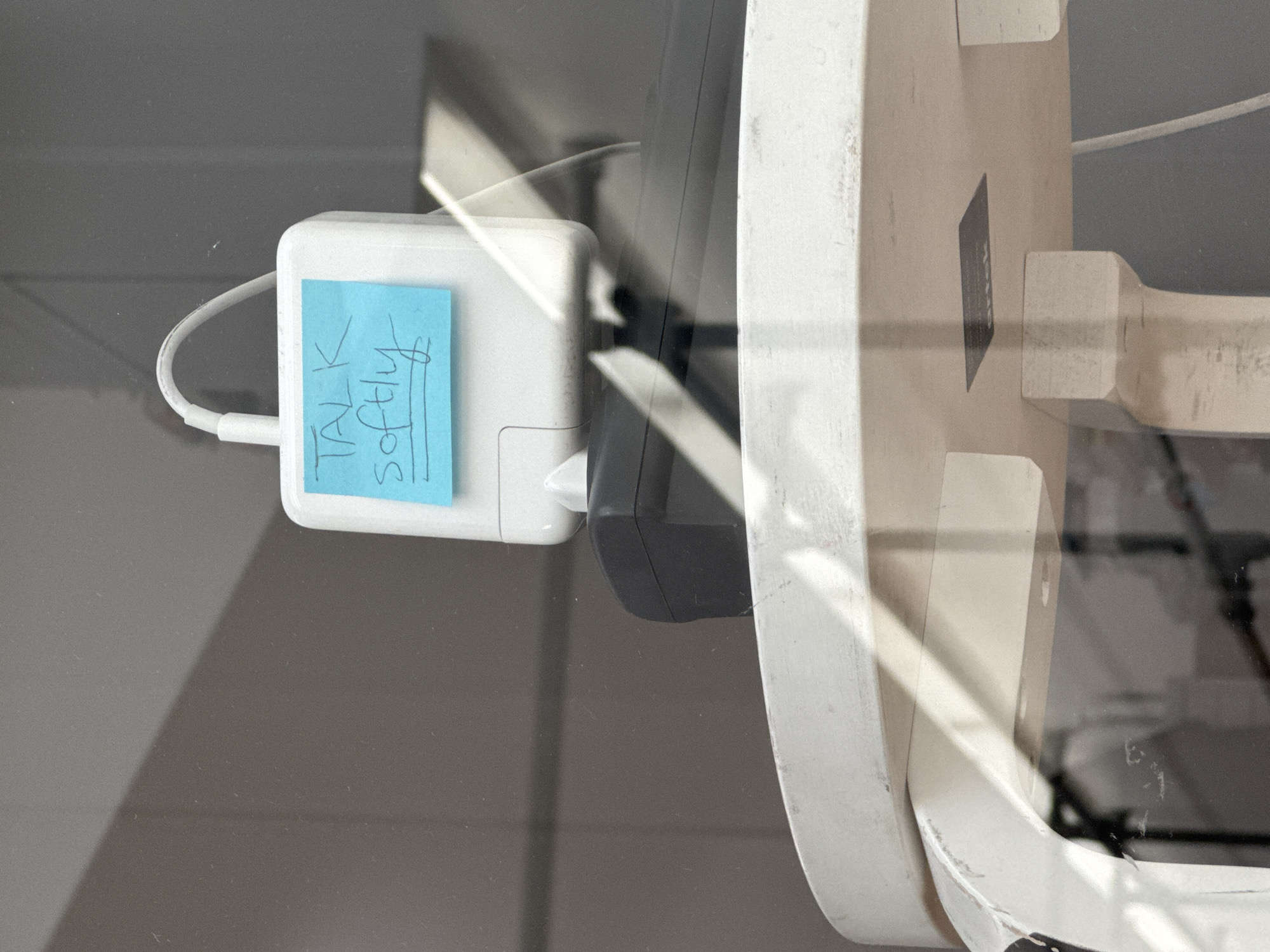 The image presents a close-up view of electronic equipment observed through a reflective surface, likely glass, which shows strong diagonal streaks of sunlight. In the central foreground, a white power adapter, consistent with an Apple MacBook charger, is visible with a white cable attached to its left side. A light blue sticky note is affixed to the side of the power adapter, displaying the handwritten text "TALK SOFTLY" in dark, capitalized letters. The adapter is plugged into a darker, possibly black, electronic component or power outlet on its right.
To the right of the central equipment, a large, off-white, and somewhat worn or dirty object, possibly a piece of furniture, a stand, or a structural element like a window frame, is partially visible. The background consists of a blurred, dark grey surface. The distinct light reflections suggest the photo was taken during bright daylight hours. No people are present, and no specific activity is occurring beyond the passive state of the plugged-in adapter. The scene appears to be an indoor setting, potentially a workspace or an area where electronics are stored or displayed behind a transparent barrier. There are no visual cues within the image itself to confirm the provided location context of Paris, France.