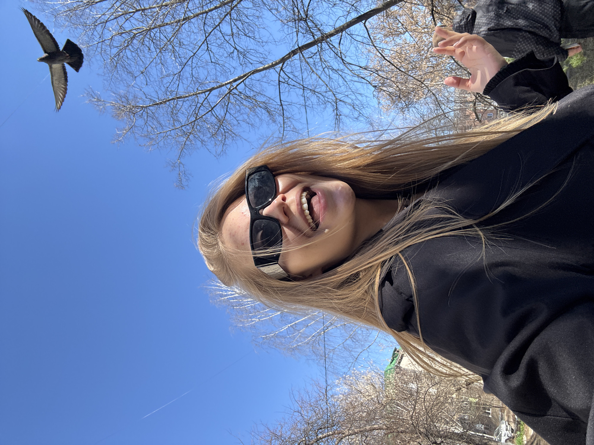 The image shows a selfie of a fair-skinned person with long blonde hair, wearing black sunglasses and a black sweatshirt. The person is smiling with their mouth open. Above and to the left, a bird is in flight against a clear blue sky. Bare tree branches are visible in the upper portion of the frame. Another person's hand, wearing a black and grey patterned jacket, is partially visible in the upper right corner. In the background, there are trees and what appears to be a building with a green roof. The location is Chaikovsky, Russia.