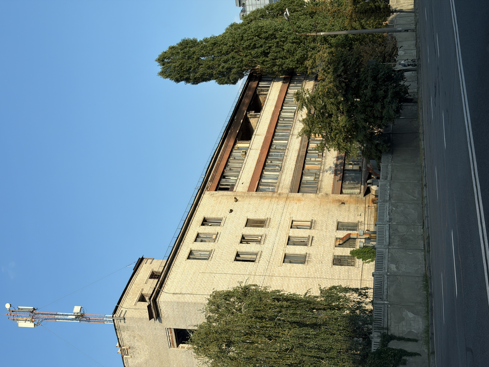 This is an exterior, eye-level, medium shot of an abandoned or under-renovation multi-story brick building in Kyiv, Ukraine. The building is rectangular and has several rows of windows, many of which are broken or boarded up. The facade is made of light-colored bricks and shows signs of age and neglect. A cell tower is visible on the roof of the building, with multiple antennas. Mature trees, one tall and slender, the other fuller and weeping, flank the building. A concrete fence runs along the front of the property, separating it from a two-lane asphalt road. The sky is clear and blue. A person on a bicycle is visible on the sidewalk behind the fence, near the tall tree. The scene suggests a post-industrial or developing urban environment.