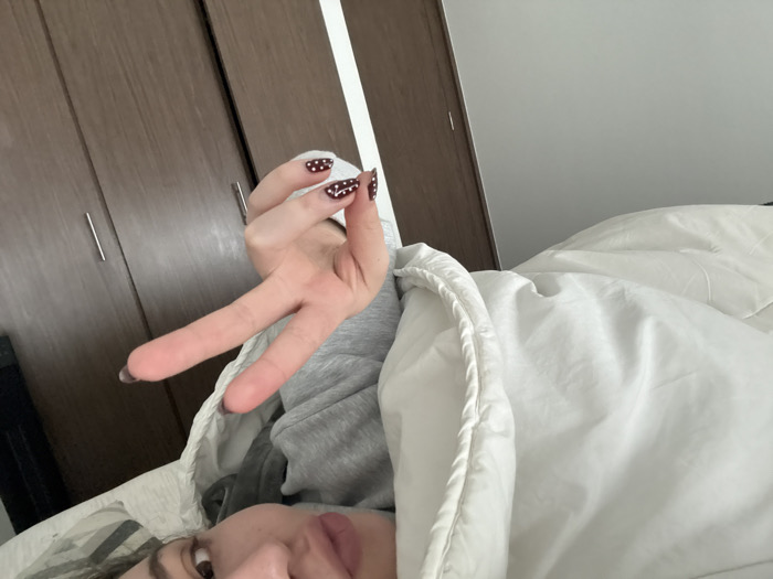 An indoor scene depicts a person lying down, viewed from a low angle, likely from a bed. The lower portion of a person's face is visible in the bottom left, featuring light pink lips, part of a brown eye, and dark, wavy hair. A right hand, positioned centrally, is extended with the index and middle fingers raised in a 'V' gesture. The fingernails are painted dark red with white polka dots; one nail also displays a white wavy line pattern. The person is partially covered by a white comforter with a prominent corded edge. A grey long-sleeved garment is visible beneath the comforter. The background consists of two closed wooden wardrobe doors with vertical silver handles on the left, and a light grey wall to the right. The specific location within the city of Mexico City, Mexico, cannot be determined from the visual evidence.