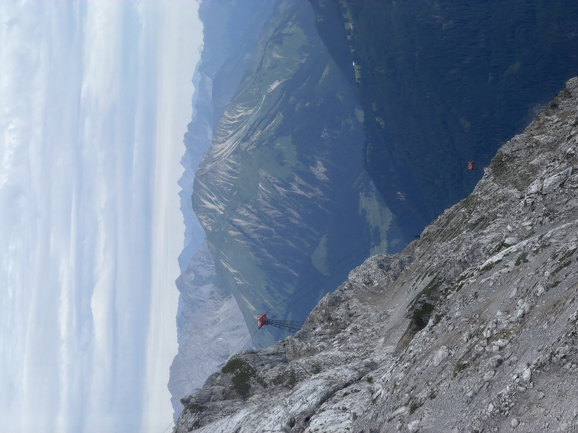 The image shows a dramatic mountain landscape with a cable car station perched on a steep, rocky incline. The cable car station is red and sits atop a dark metal support structure. Cables extend from the station across the valley, connecting to unseen points. The foreground is dominated by a sheer cliff face covered in scree and small patches of vegetation. In the background, layers of green, tree-covered mountains recede into the distance under a cloudy sky. The overall scene is one of rugged natural beauty and man-made infrastructure in a high-altitude environment. This location is likely in the vicinity of Grainau, Germany, given the presence of cable cars in mountainous regions surrounding the town, such as the Zugspitze railway.