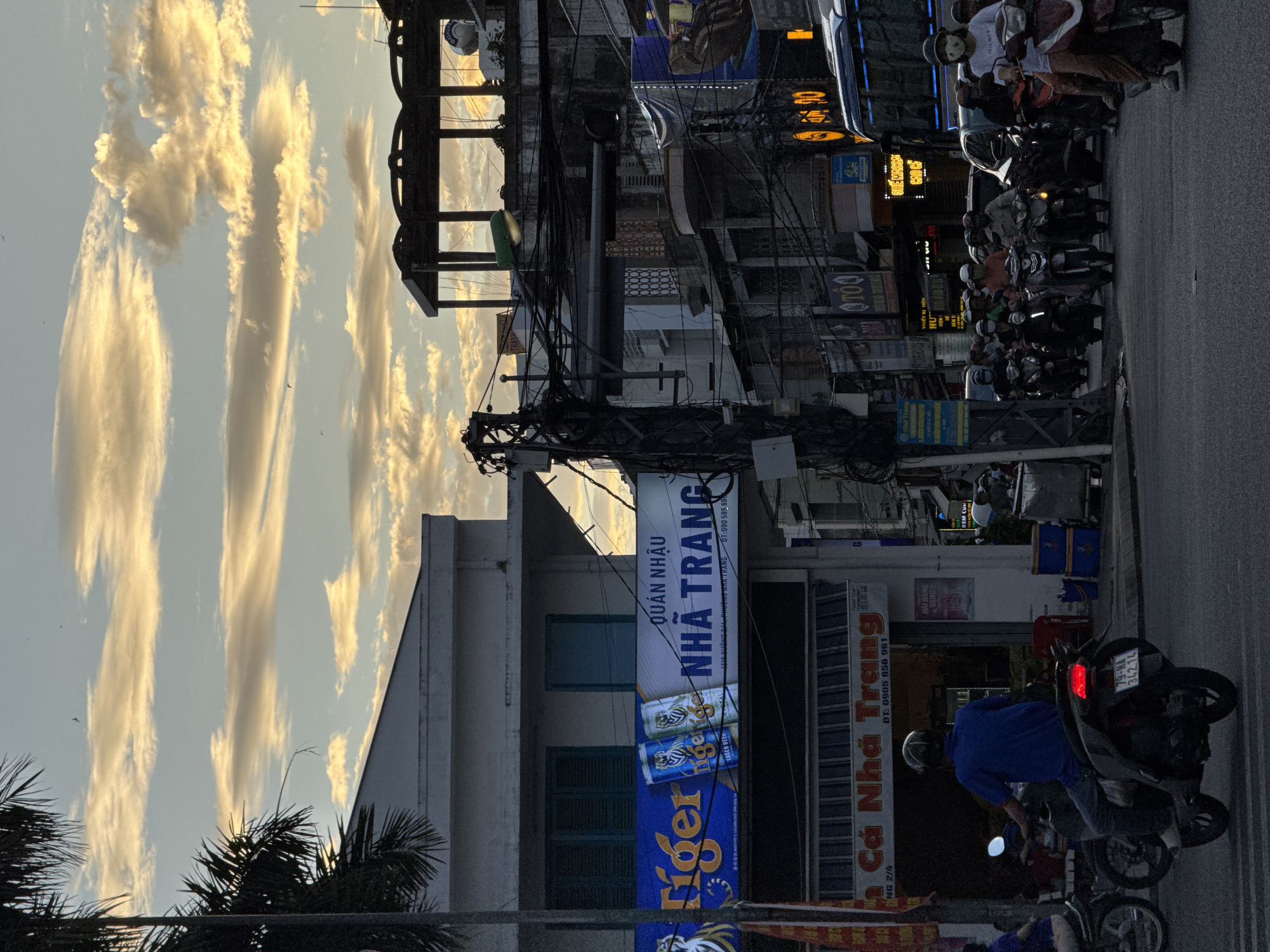 The image depicts a street scene in Nha Trang, Vietnam, likely during the late afternoon or early evening, given the golden hues in the sky and the presence of artificial lights. The sky is filled with scattered clouds, some illuminated by the setting sun.
In the foreground, a person wearing a blue shirt and dark pants is riding a scooter, facing away from the camera. The scooter has a license plate that reads "79-MA 342.14". Behind this scooter, other motorcycles and scooters are visible, with many riders wearing helmets, some with masks. To the left, another scooter and rider are partially visible.
Buildings line the street. One building features a prominent blue and yellow sign with "Tiger" branding and cans of Tiger beer depicted. Adjacent to this is a sign with Vietnamese text, including "QUÁN NHẬU NHÀ TRANG" (Nha Trang Bar/Restaurant). Another sign below reads "CÁ NHÀ TRANG" (Nha Trang Fish).
The street is filled with a dense network of electrical wires, characteristic of many Vietnamese cities. Further down the street, a bus is visible, and more buildings with various signs and storefronts can be seen. The overall impression is a busy urban environment with significant traffic and a vibrant street life.