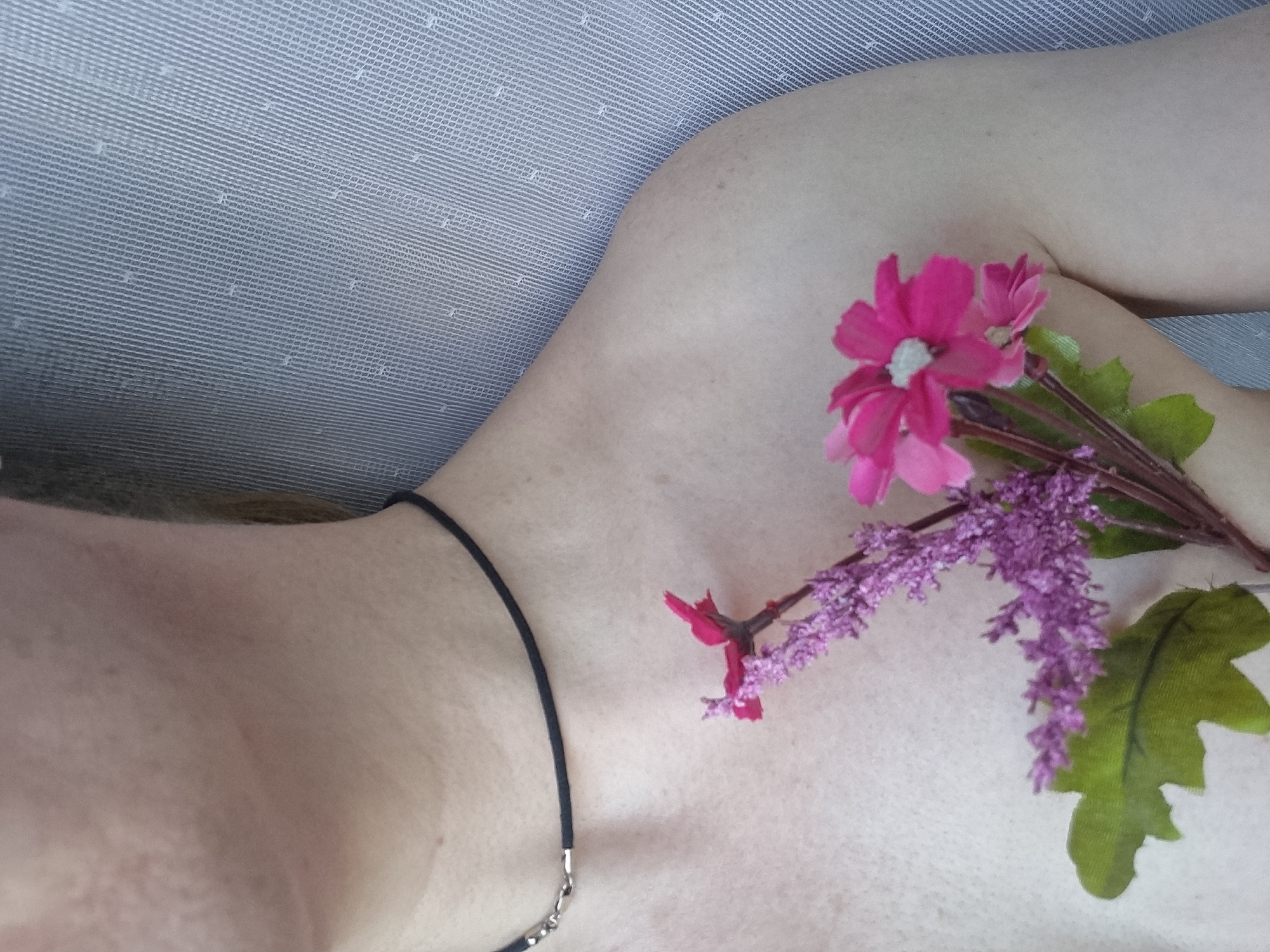 The image presents a close-up, high-angle view of a person's upper torso, focusing on the shoulder, clavicle, and upper chest area. The skin, appearing fair, shows some minor blemishes and faint freckles. A thin black cord necklace with a small silver clasp is worn around the neck, extending into the frame from the left.
A small arrangement of artificial flowers is positioned on the person's right shoulder and upper chest. This floral cluster includes prominent bright pink flowers with white centers and green leaves, alongside more delicate sprigs of light purple flowers.
The background is a light grey or off-white textured surface, possibly fabric or a mesh screen, which fills the upper and left portions of the frame. The overall composition suggests a relaxed or artistic pose, with the flowers placed decoratively on the body.
No specific activity is taking place beyond the static arrangement. The time of day and weather are not discernible, and there are no strong environmental cues to indicate a precise location within Chaikovsky, Russia, other than the context provided. No text is visible in the image.