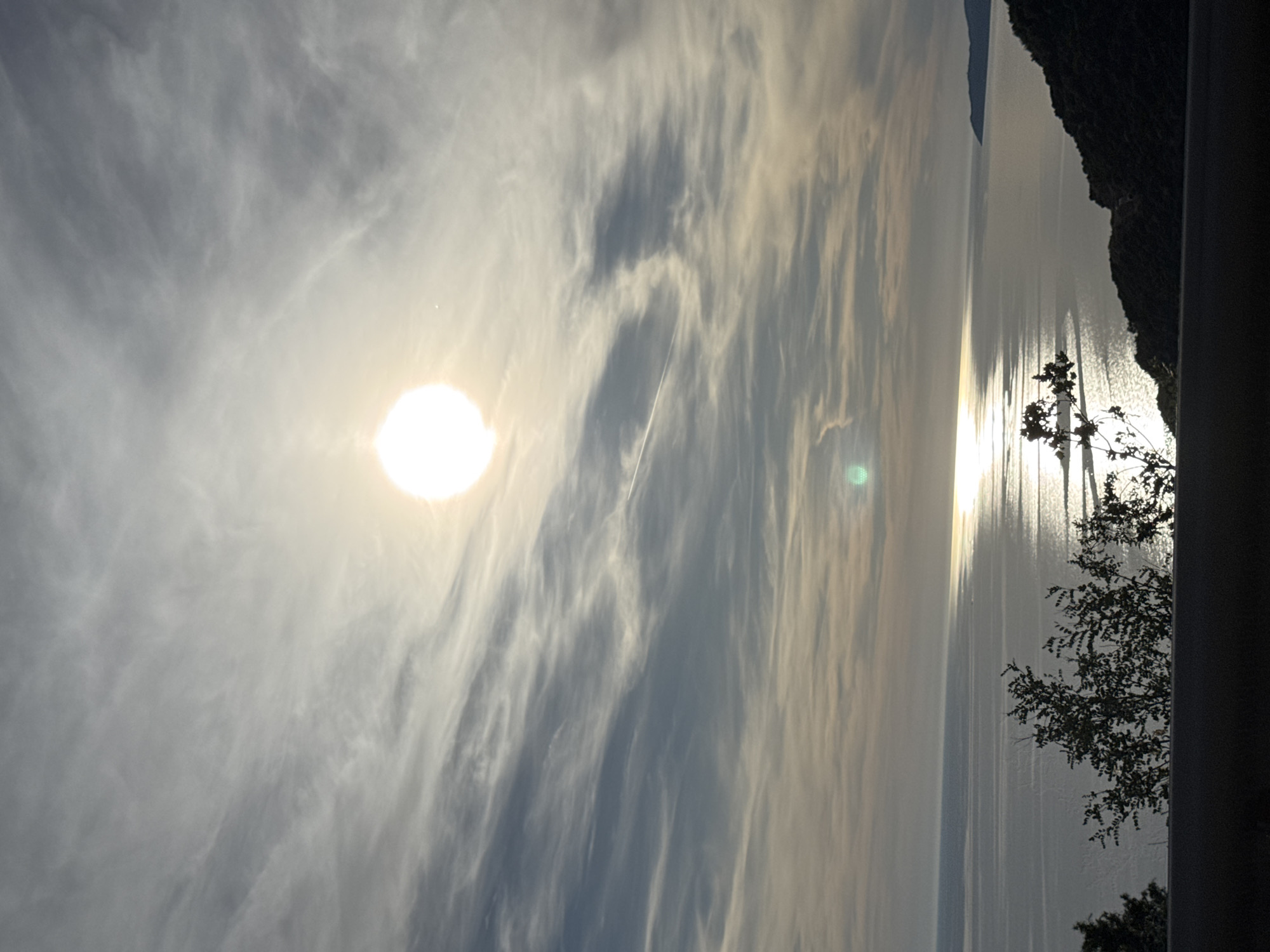 The image depicts a serene coastal landscape bathed in the warm glow of the setting sun. The sky is a canvas of wispy clouds, streaked with hues of grey, white, and pale blue, creating a dramatic backdrop for the brilliant, orb-like sun positioned in the upper center of the frame. The sun's reflection dances upon the calm, rippling surface of the sea, forming a shimmering pathway of light that stretches towards the horizon.
In the foreground, the dark silhouette of a hill covered in lush, green vegetation slopes down towards the water on the right side of the image. A sparse, dark bush with delicate branches and leaves stands prominently in the lower left, its silhouette stark against the luminous sea. The lower edge of the frame is occupied by a dark, horizontal band, possibly a road or embankment, from which the viewpoint is taken.
The scene is devoid of any human presence or activity, emphasizing the natural beauty and tranquility of the location. The overall impression is one of peacefulness and a quiet appreciation of the natural world, characteristic of coastal vistas. The image captures a moment of striking natural beauty, where the interplay of light and shadow on the sky, sea, and land creates a captivating panorama.