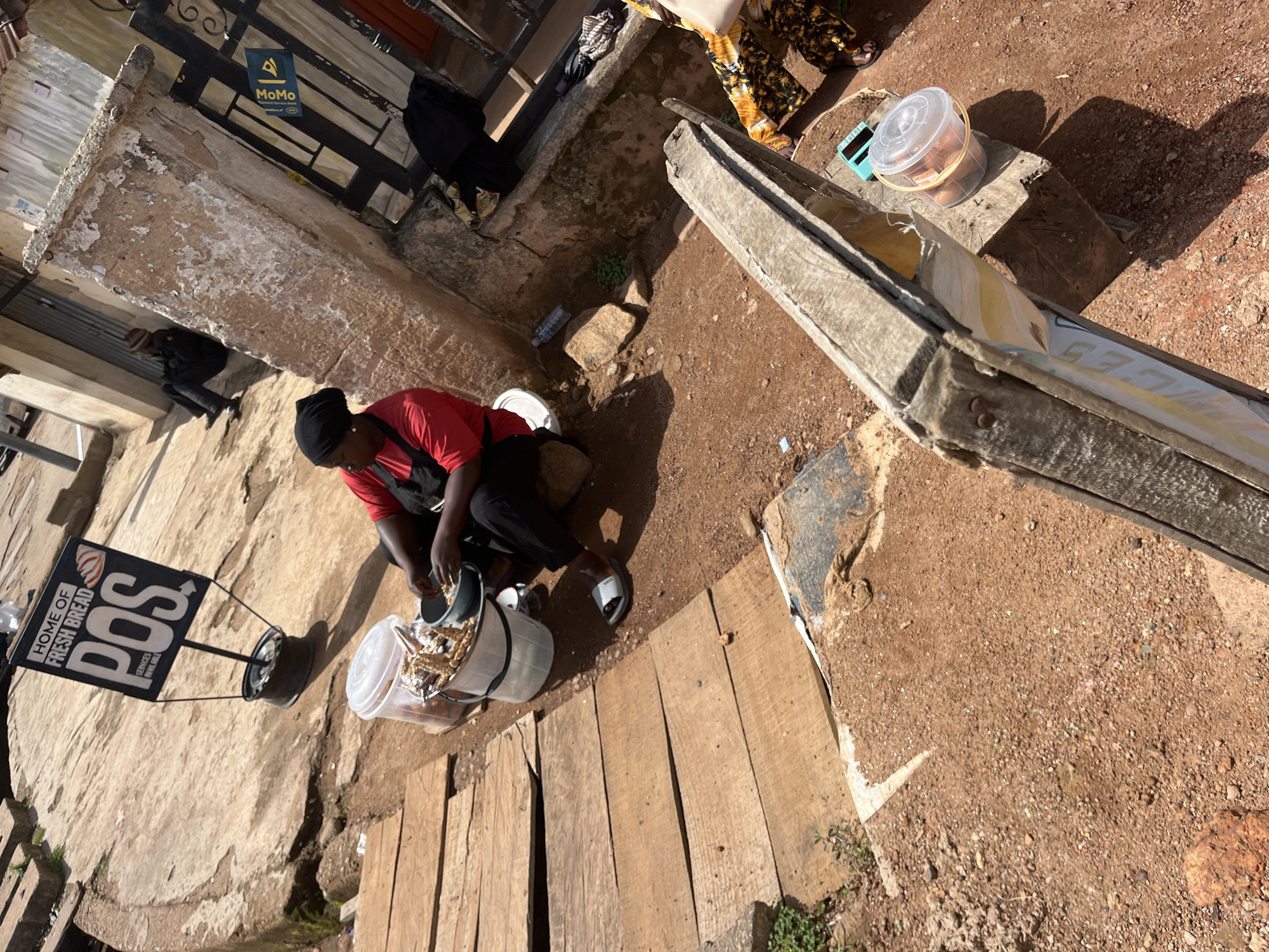 A woman wearing a red shirt and black headscarf is preparing food in Gura Top, Nigeria. She is crouched next to a white bucket filled with snacks and a green bowl containing some food items. The scene is set on a dirt ground with wooden planks forming a makeshift pathway. Behind her, a sign advertises "HOME OF FRESH BREAD POS SERVICES AVAILABLE". In the background, a man sits on a ledge near a building with a metal door, and a blue Momo sign is visible on the door. The environment appears to be an outdoor market or street vendor setting with scattered debris and a few bottles on the ground.