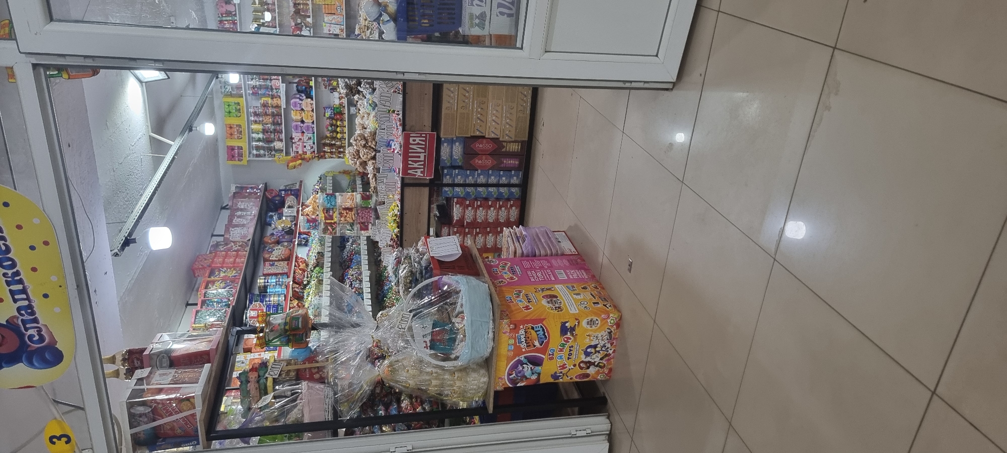 The image depicts the interior of a small shop selling candy and toys. The shop is filled with shelves stocked with various packaged sweets and boxes of toys. In the foreground, there are display baskets filled with gift-wrapped items and what appear to be toy gift sets. A sign with the word "АКЦИЯ!" (meaning "SALE!" in Russian) is visible on a shelf in the middle of the image. The flooring is tiled. The overall impression is of a retail environment characterized by abundance of merchandise, primarily focused on confectionery and children's toys. The shop is located in the city of Aktobe, Kazakhstan.