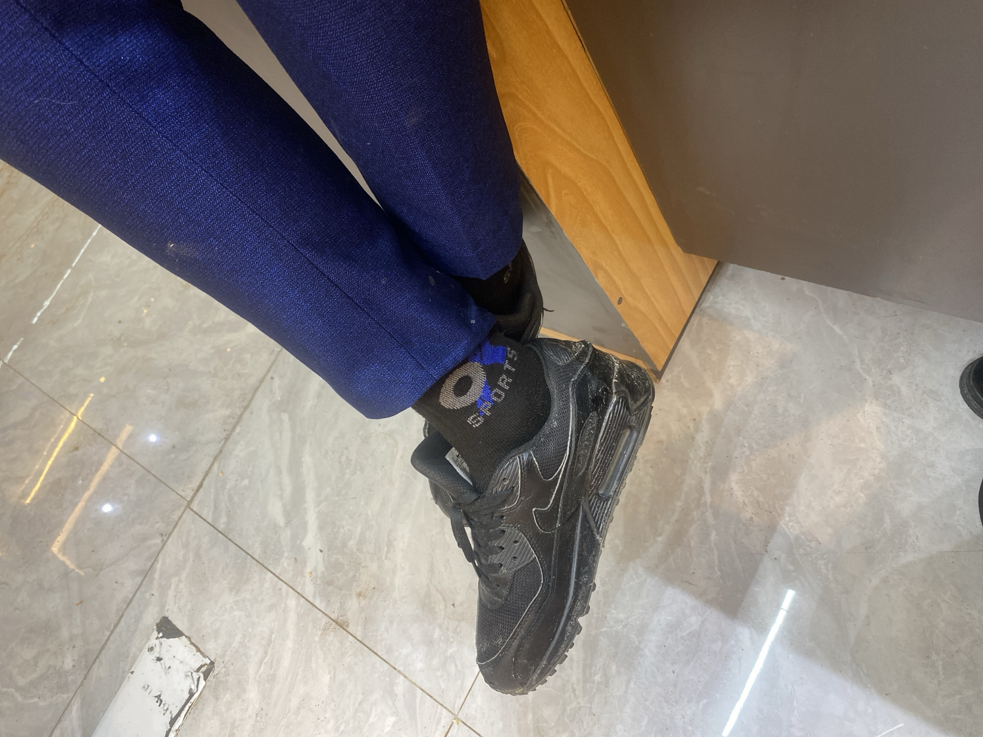 A low-angle shot features the lower portion of a person sitting, wearing dark blue dress pants, black socks with "SPORTS" text, and black Nike Air Max 90 sneakers. The person's right leg is extended towards the lower right of the frame, while the left leg is slightly retracted. The setting appears to be indoors, with light-colored marble flooring. A light wood panel and dark wall are visible in the upper right. The scene does not provide enough information to determine if the location is in Dar es-Salaam, Tanzania.