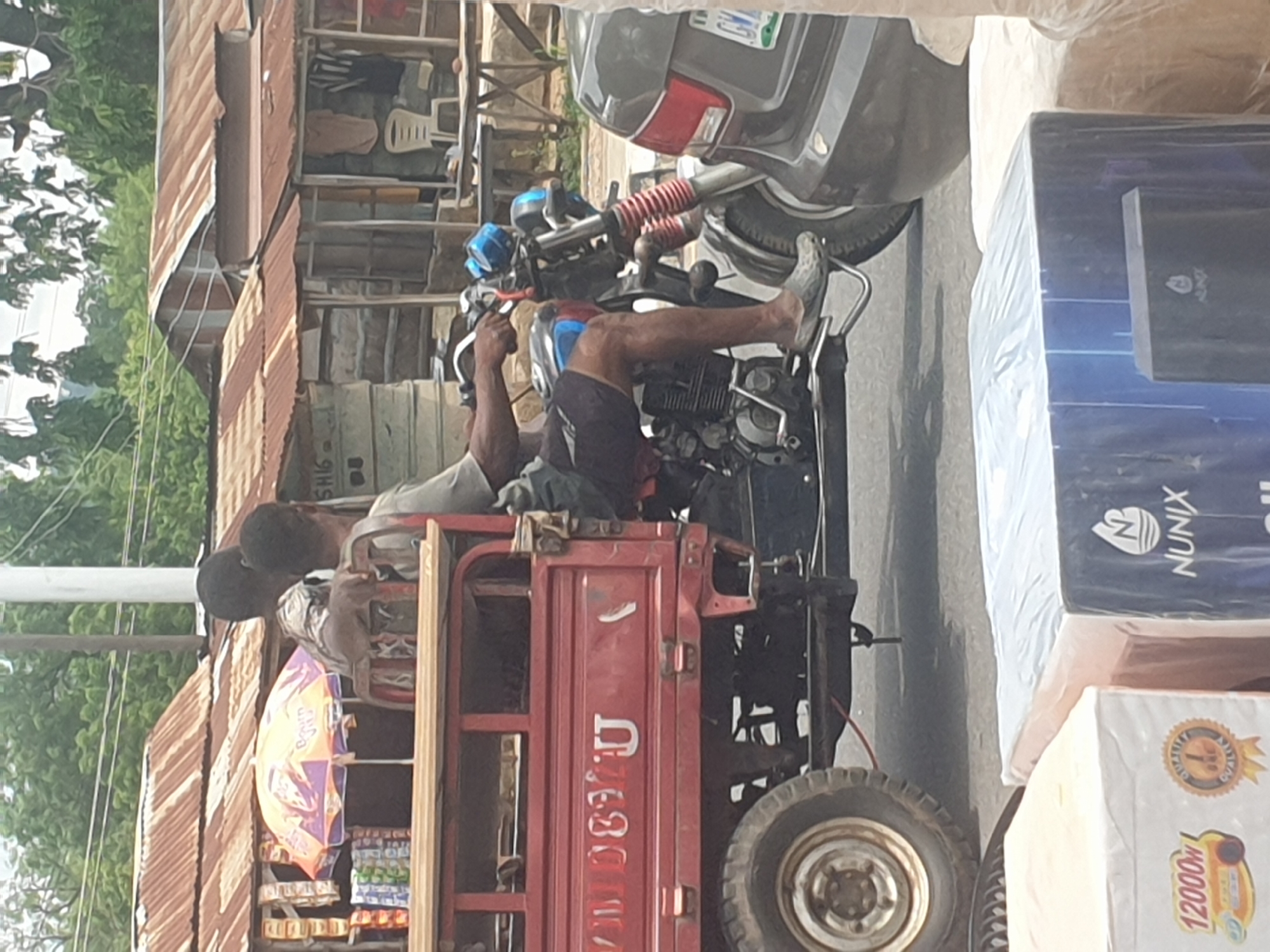 In Adankolo, Nigeria, a three-wheeled motorcycle, commonly used for transport, stands on the road. The red vehicle is loaded with goods, including boxes of products and a display of snacks and sweets under an umbrella. Two men are seated on the vehicle. A motorcycle is parked next to it. A dark grey car is nearby. Behind the vehicle, corrugated iron roofs top buildings, interspersed with trees. Packaged goods are stacked beside the vehicle.