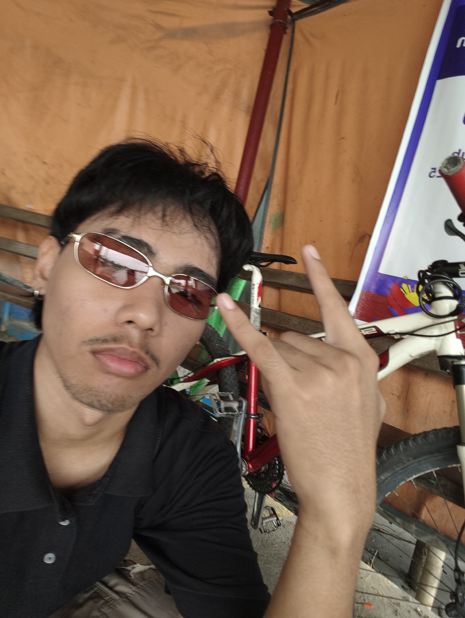 The media file is a selfie of a young man wearing sunglasses and a black polo shirt. He is making a peace sign with his right hand. The background appears to be outdoors, with a large orange tarp or fabric. Behind the man, parts of a bicycle are visible, including a red frame and a white frame. There's also a tall, vertical banner or sign with blue and white trim. The lighting suggests it could be daytime. The man has dark hair and a light mustache. There is some stubble on his chin and jawline. He is wearing a silver hoop earring in his left ear. The overall impression is casual and informal.