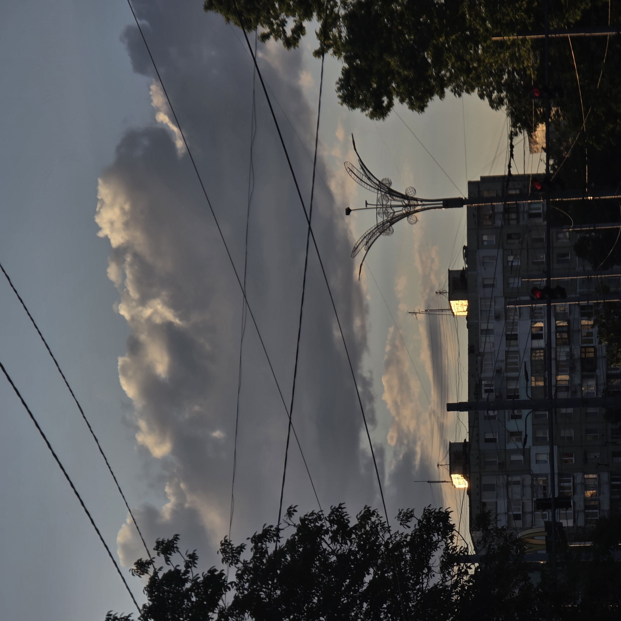 The image depicts an urban scene at dusk, dominated by a large, multi-story residential building. The sky is filled with a mix of dark and light clouds, with patches of soft, warm light suggesting the setting sun. Several power lines crisscross the frame, creating a layered effect against the sky.
The building itself appears to be constructed from concrete and has numerous windows, many of which reflect the ambient light. On the roof of the building, a smaller structure is visible, catching a bright reflection of sunlight. To the left of the main building, a silhouetted tree with dense foliage is present. In the foreground, a streetlamp with an ornate, decorative top is visible on the right side of the image. A traffic light with a red illuminated signal is positioned below the streetlamp. A portion of a McDonald's sign is also subtly visible on the left side of the building. The overall atmosphere is one of an ordinary urban evening.