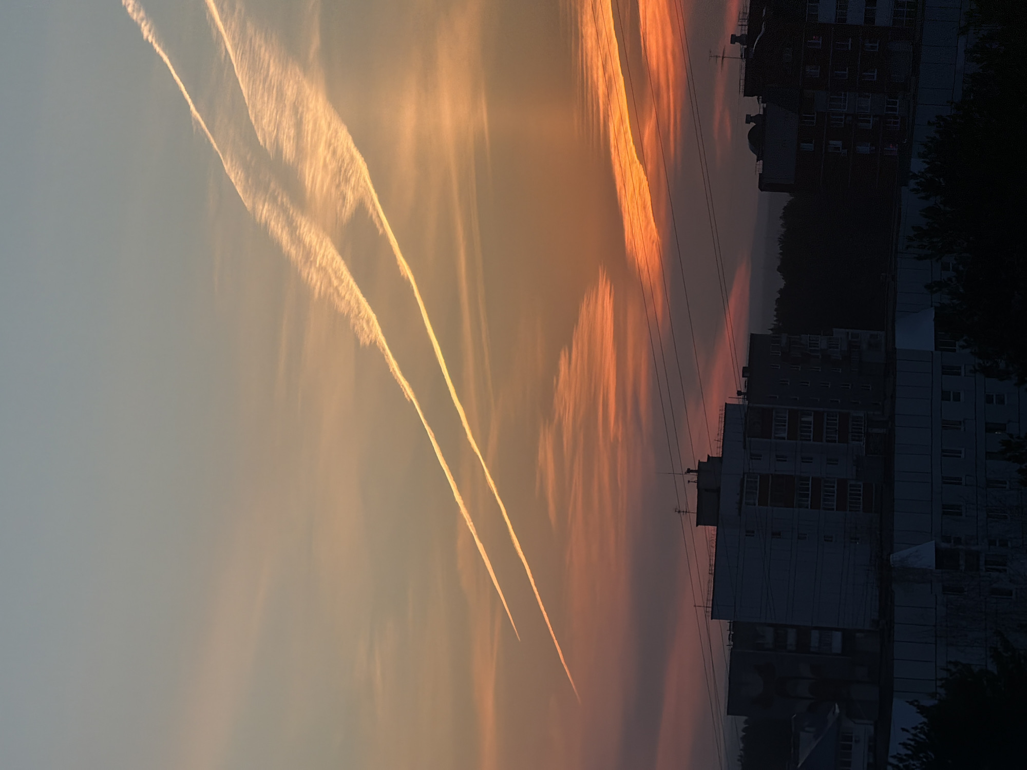 The image depicts a sky with contrails and orange hues, suggestive of a sunset or sunrise. In the background, there are buildings, likely residential, and some trees. Power lines are also visible stretching across the sky. Based on the prompt's requirement for a location in Chaikovsky, Russia, this image could be a residential area in that city. There are no people visible in the image. The scene captures the intersection of natural atmospheric phenomena (contrails, sky coloration) with the built environment of a city.