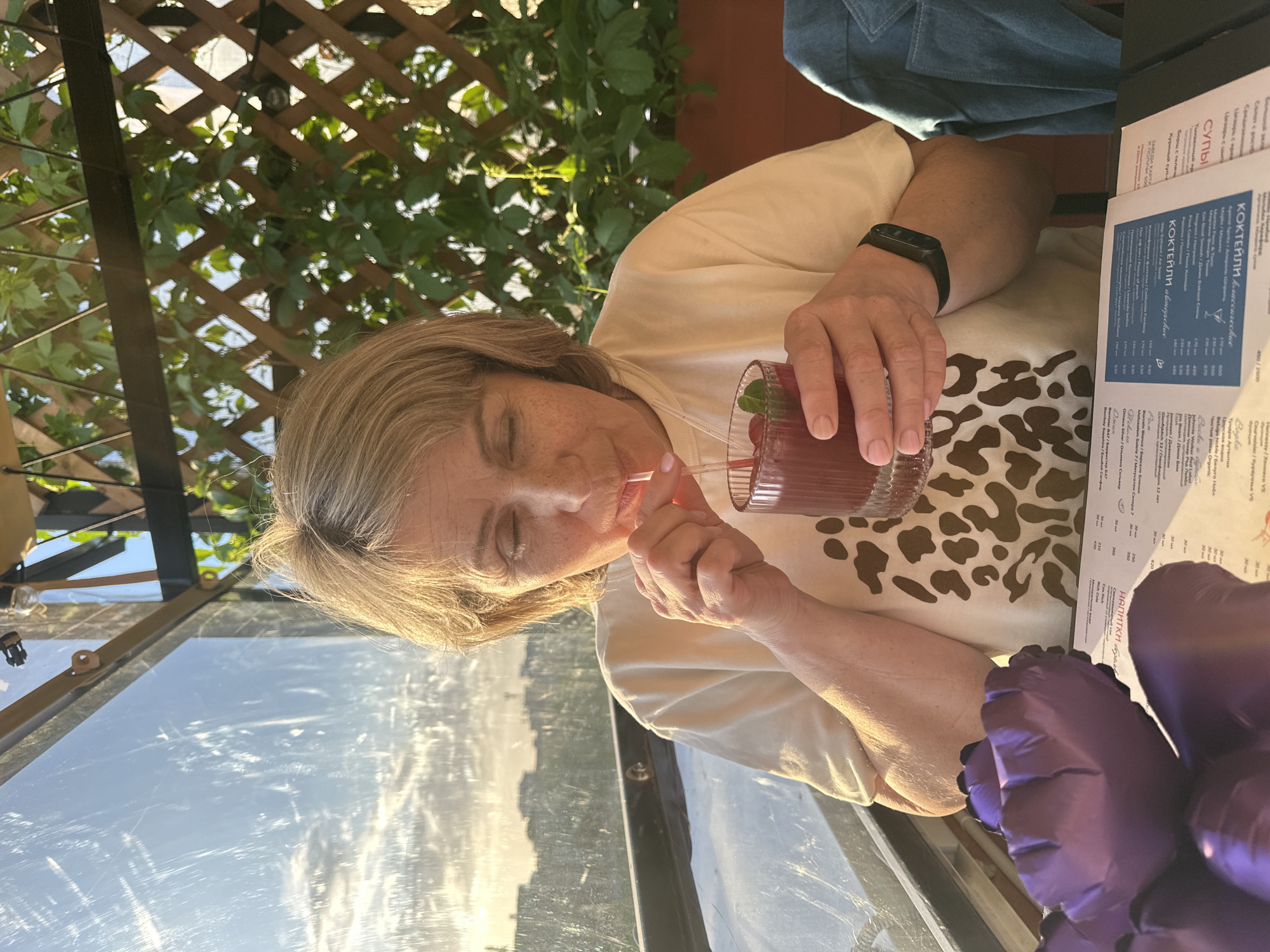 The photograph shows a blonde woman seated at a table outdoors, possibly at a restaurant or cafe, as there is a menu visible in front of her. She is drinking a red cocktail with a straw and mint garnish. A partially visible trellis with foliage suggests a patio setting. The photo appears to be taken from a slightly tilted angle and shows an outdoor view in the background, suggesting the location is a city, possibly Perm, Russia due to the Cyrillic menu with Russian text. Another individual wearing a denim jacket is partly visible in the upper right corner, and a pair of purple gloves is in the bottom right of the frame.