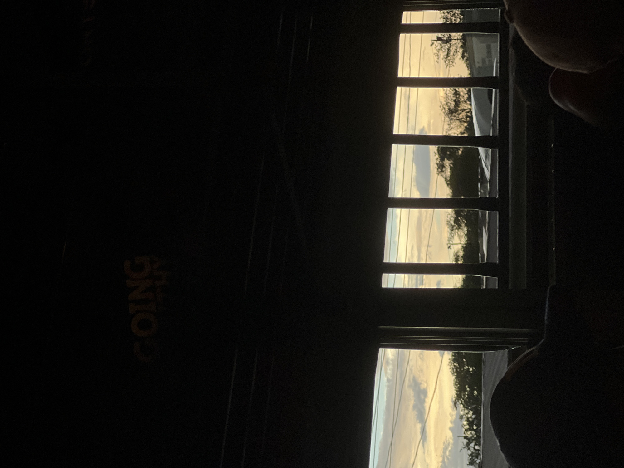 The image depicts a view from an interior space looking out through a window or opening at a sunset. The foreground is dominated by dark, silhouetted shapes, likely furniture or people, suggesting the viewer is inside a building or vehicle. Above these shadows, the window reveals a sky with a gradient of warm colors, transitioning from a pale blue and white with scattered clouds to a strong yellow-orange hue near the horizon. Silhouetted trees and structures are visible against this sky. The window is framed by dark, vertical bars, creating a grid-like effect over the outdoor scene. In the upper part of the image, within the interior darkness, there are yellow illuminated letters that read "GOING". The scene appears to be set in El Guasaule, Nicaragua, as indicated by the prompt's context.
