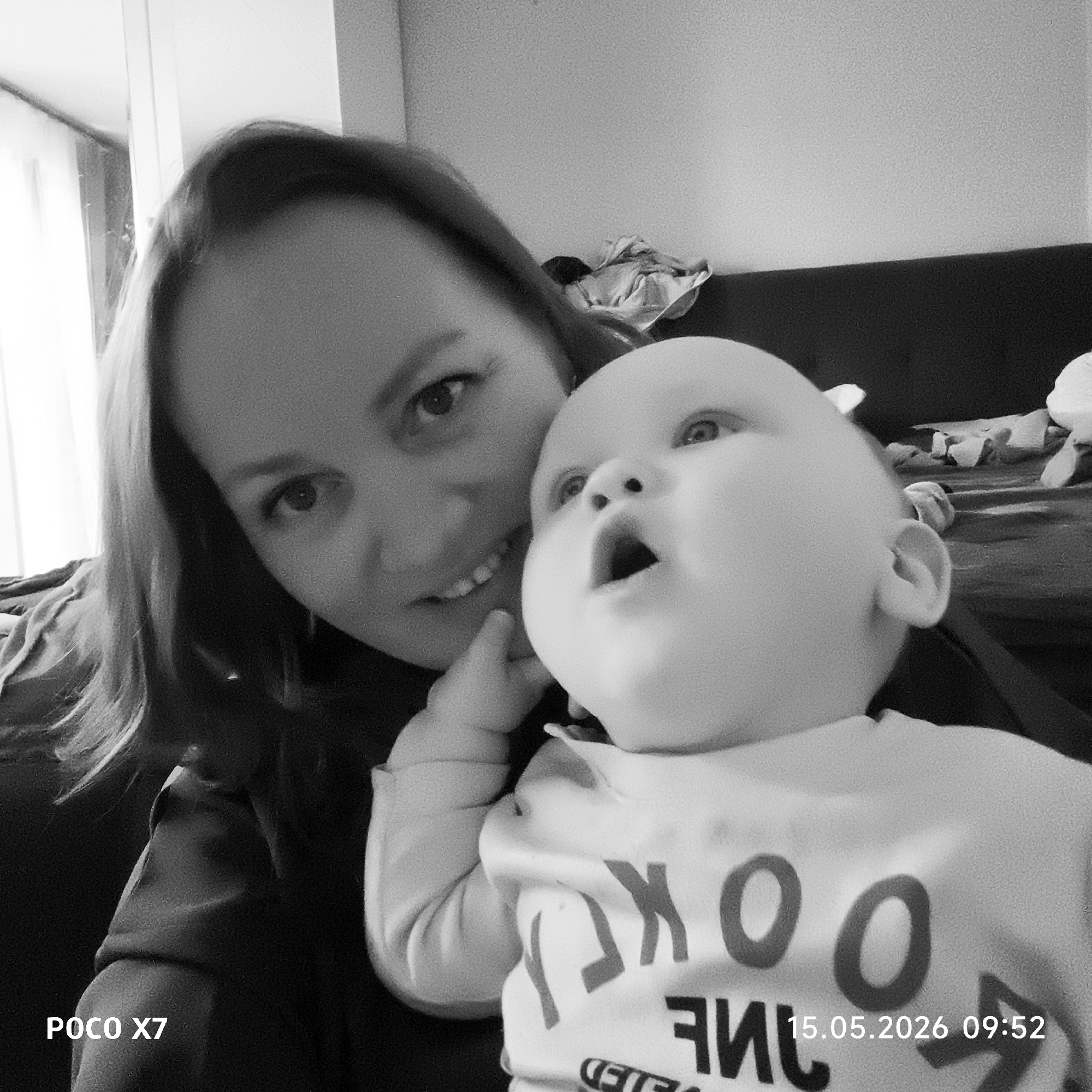 A woman and a baby are captured in a black and white photograph. The woman is on the left side of the frame, smiling and looking at the camera. The baby is positioned in front of her, gazing upwards with their mouth open. The baby is wearing a light-colored shirt with reversed text. The background includes a dark couch and some indistinct objects. The text "POCO X7" is visible in the bottom left corner, and the date "15.05.2026 09:52" is in the bottom right corner. The location is indicated as Chaikovsky, Russia. The subjects are interacting through proximity.