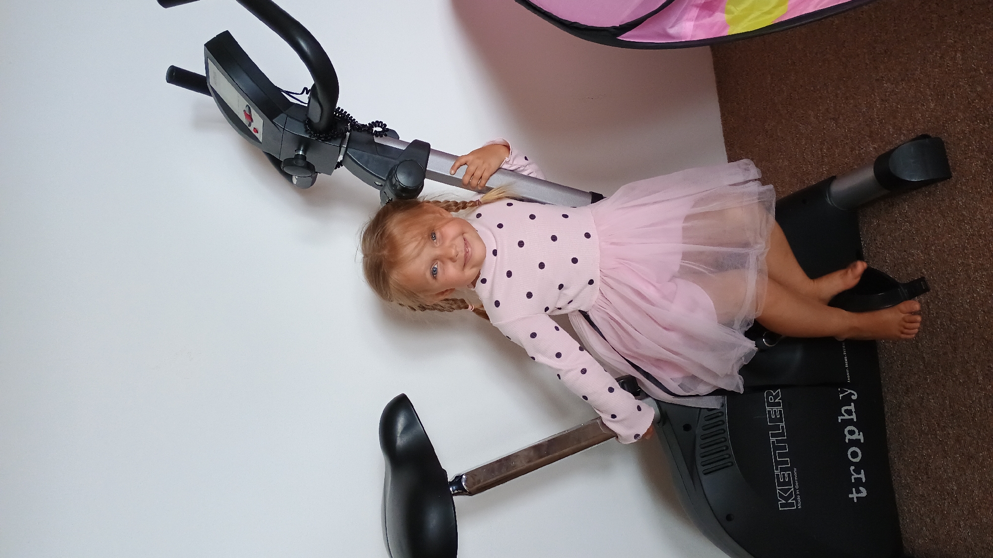 In a room with beige carpet and a white wall, a young Caucasian girl with blonde hair in pigtails sits on a black Kettler stationary bike. She is wearing a pink long-sleeved shirt with dark polka dots and a pink tutu. Her feet are bare and resting on the bike's pedals. She is smiling at the camera and holding onto the bike's central support pole. The stationary bike has a black seat, a digital display screen, and black handlebars. To the right of the bike, a pink tent with yellow accents is partially visible. The overall scene is indoors.