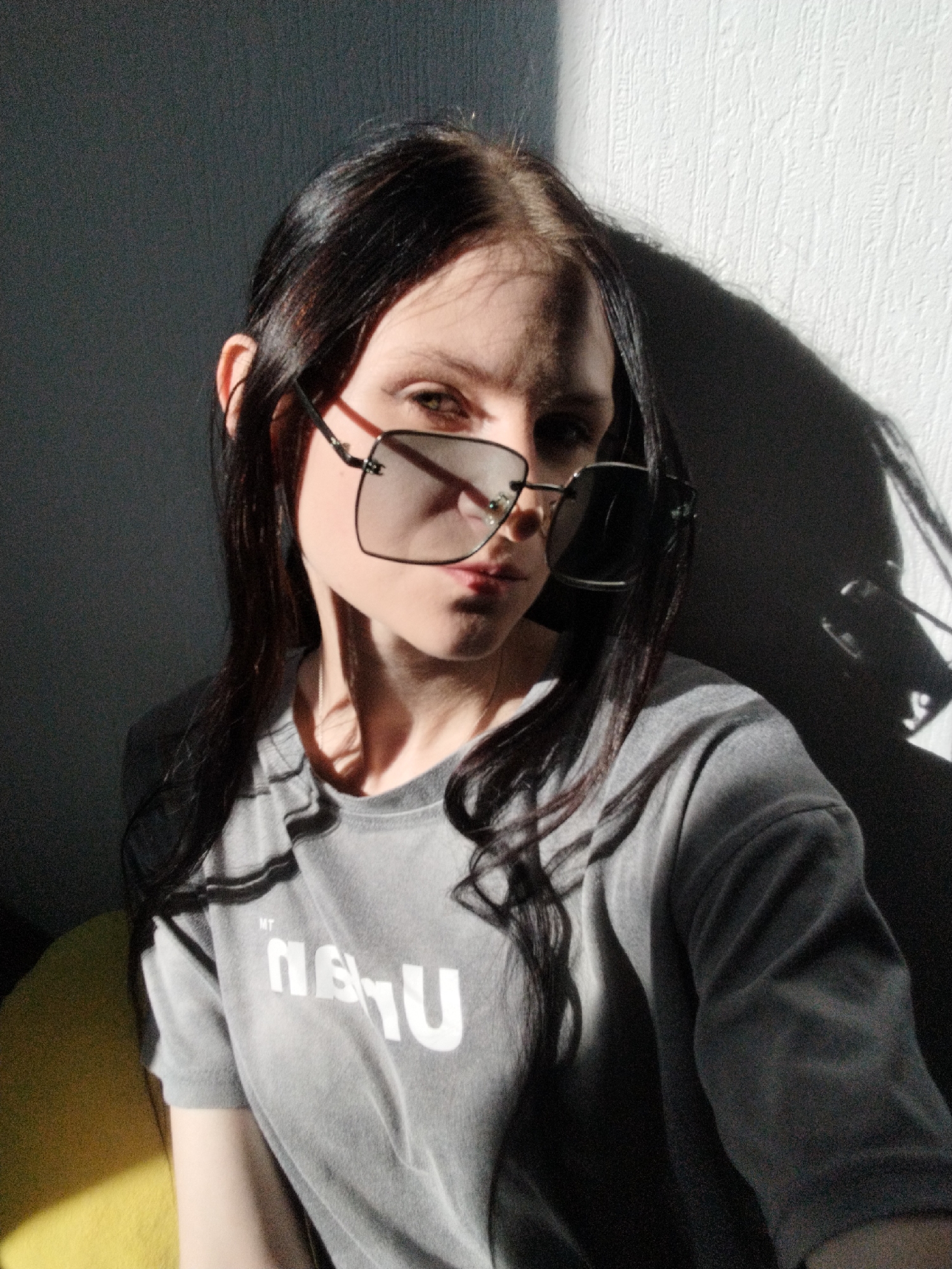This is a close-up, waist-up media file of a young woman indoors, likely in a room with a textured wall. She is wearing a gray t-shirt with white text that appears to read "MT NEU" in a stylized font. She is also wearing large, square, dark-lensed sunglasses. Her long, dark hair is visible, some of it falling over her shoulders and in front of her face.
The lighting suggests it is daytime, with strong sunlight casting dramatic shadows on the wall behind her and on her face. The harsh light highlights her features and creates a high-contrast image. Her expression, partially obscured by the sunglasses, seems to be one of contemplation or a slight pose. The overall mood is somewhat moody and atmospheric due to the lighting conditions. A hint of a yellow object is visible in the lower left corner, possibly a cushion or piece of furniture, adding a small splash of color to the otherwise muted tones. There are no explicit indicators of the specific location or the weather outside.
