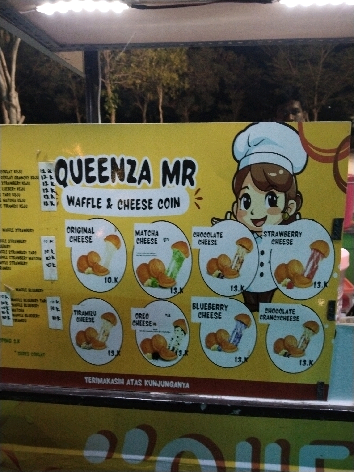This is a horizontal, medium shot image of a food stall in Slawi, Indonesia. The stall displays a yellow sign with "QUEENZA MR" prominently written in large, black, stylized letters. Below this, it says "WAFFLE & CHEESE COIN" in smaller, white text. The sign features a cartoon drawing of a girl wearing a chef's hat.
The sign lists various waffle and cheese coin flavors with accompanying prices, some with small images of the food items. Flavors visible include:
* Original Cheese (10.K)
* Matcha Cheese (13.K)
* Chocolate Cheese (13.K)
* Strawberry Cheese (13.K)
* Tiramizu Cheese (13.K)
* Oreo Cheese (13.K)
* Blueberry Cheese (13.K)
* Chocolate Cranchycheese (13.K)
There are also lists of ingredients and toppings on the left side of the sign, with prices like "12.K", "13.K", and "14.K". At the bottom of the sign, it says "TERIMAKASIH ATAS KUNJUNGANNYA", which translates to "Thank you for your visit".
The background shows trees and a slightly out-of-focus person standing behind the stall. The stall appears to be set up outdoors, possibly at a night market or street food location, given the lighting.