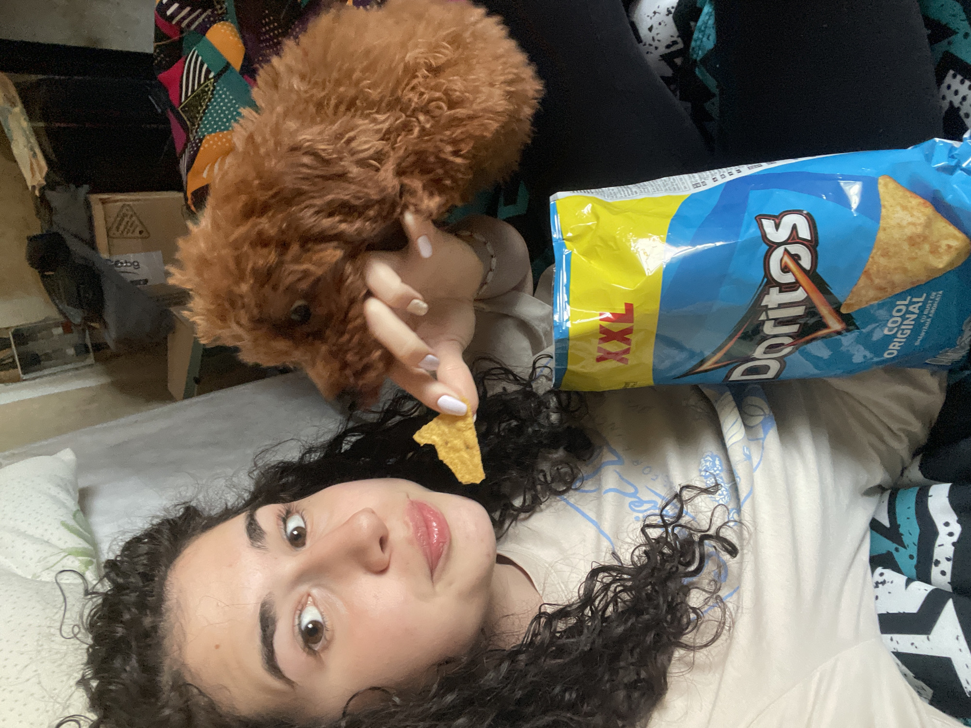 A person with curly dark hair is lying down, holding a Doritos chip in their right hand. A small brown poodle is positioned above the person's head, near the bag of Doritos Cool Original XXL. The setting appears to be an indoor space, possibly a bedroom. There is no information in the image to suggest that this photo was taken in Sofia, Bulgaria.