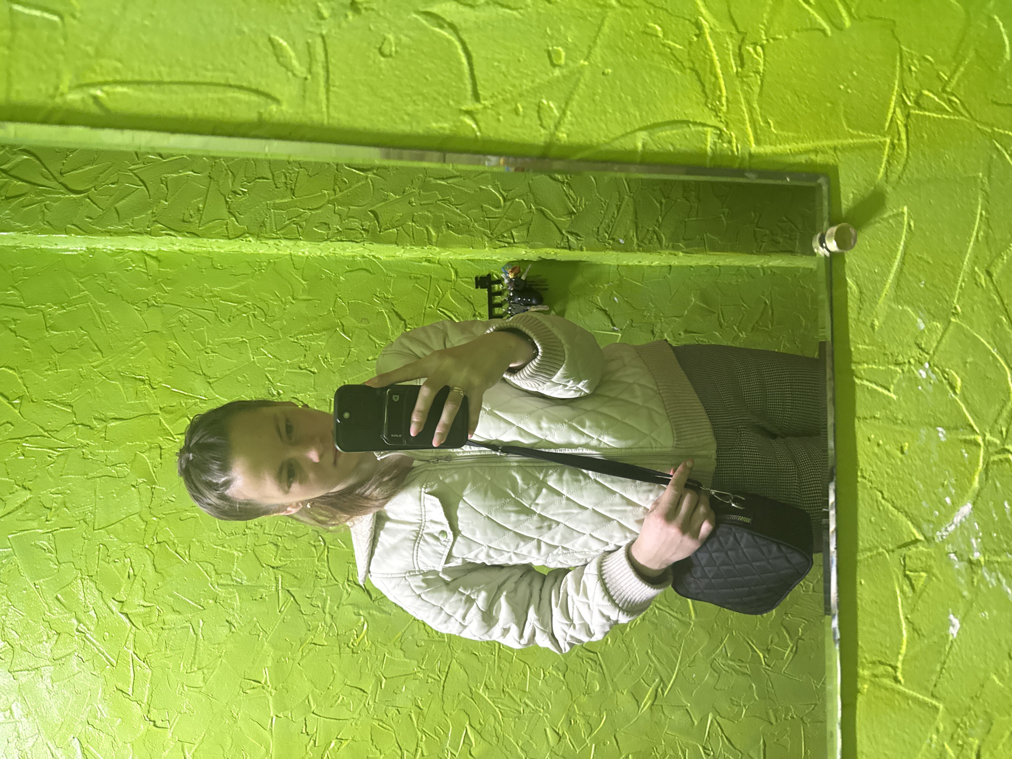 The image is a selfie taken in a mirror in a room with bright green walls. The walls have a textured finish. The mirror is rectangular with a silver frame and is attached to the wall. A person is reflected in the mirror, holding a black smartphone in their left hand and pointing it at the mirror. They are wearing a white quilted jacket and patterned pants. A black quilted purse hangs from their shoulder. A small decorative figure is visible on the wall shelf above the mirror. The location is Chaikovsky, Russia.
