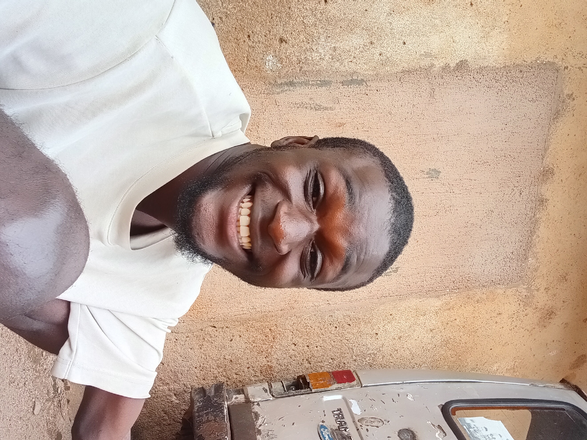 In Bukuru, Nigeria, a man with dark skin and a short haircut smiles broadly, showing his teeth. He wears a light-colored t-shirt. The background consists of a textured, light brown wall. To the right of the man, a portion of a light-colored vehicle is visible, with "TRANSIT" and "Ford" branding on its rear. The vehicle appears weathered, with visible rust and damage. The ground beneath the man and vehicle is composed of dirt and small stones. The overall scene suggests an outdoor setting in a non-urban area.