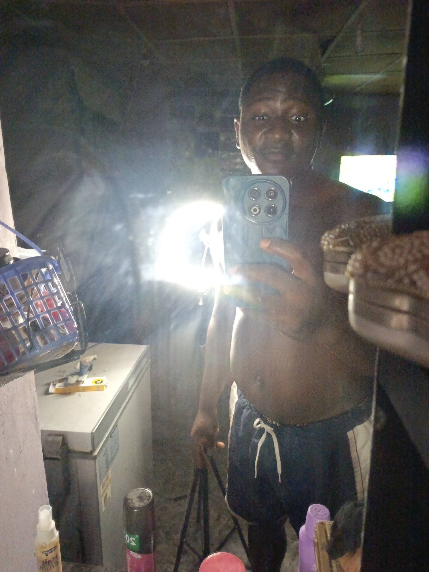 A shirtless man in dark shorts takes a selfie in a room with a refrigerator, cleaning supplies, and other miscellaneous items. The reflection in the mirror makes the lighting appear harsh, and the room's location is Unknown, Unknown.