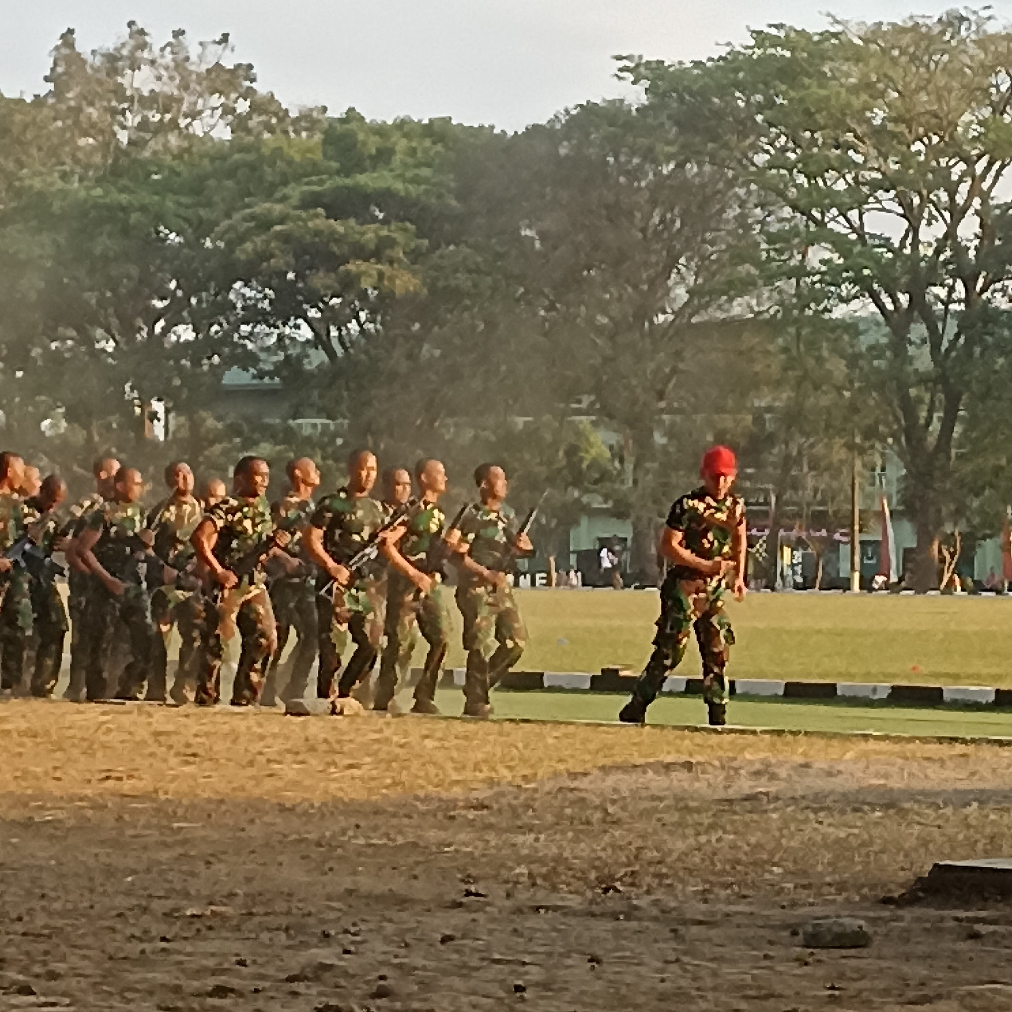 A group of soldiers in camouflage uniforms with rifles are marching in formation on a dirt field in front of a grassy area with trees in the background. One soldier in the foreground on the right, wearing a red cap, is walking in a different direction than the others. The scene appears to be an outdoor military training exercise or parade. The location in the city of Magelang, Indonesia is likely within a military academy or training ground, potentially at the Akademi Militer (AKMIL), given the presence of uniformed personnel and marching drills. The time of day appears to be early morning or late afternoon, with soft, diffused lighting.