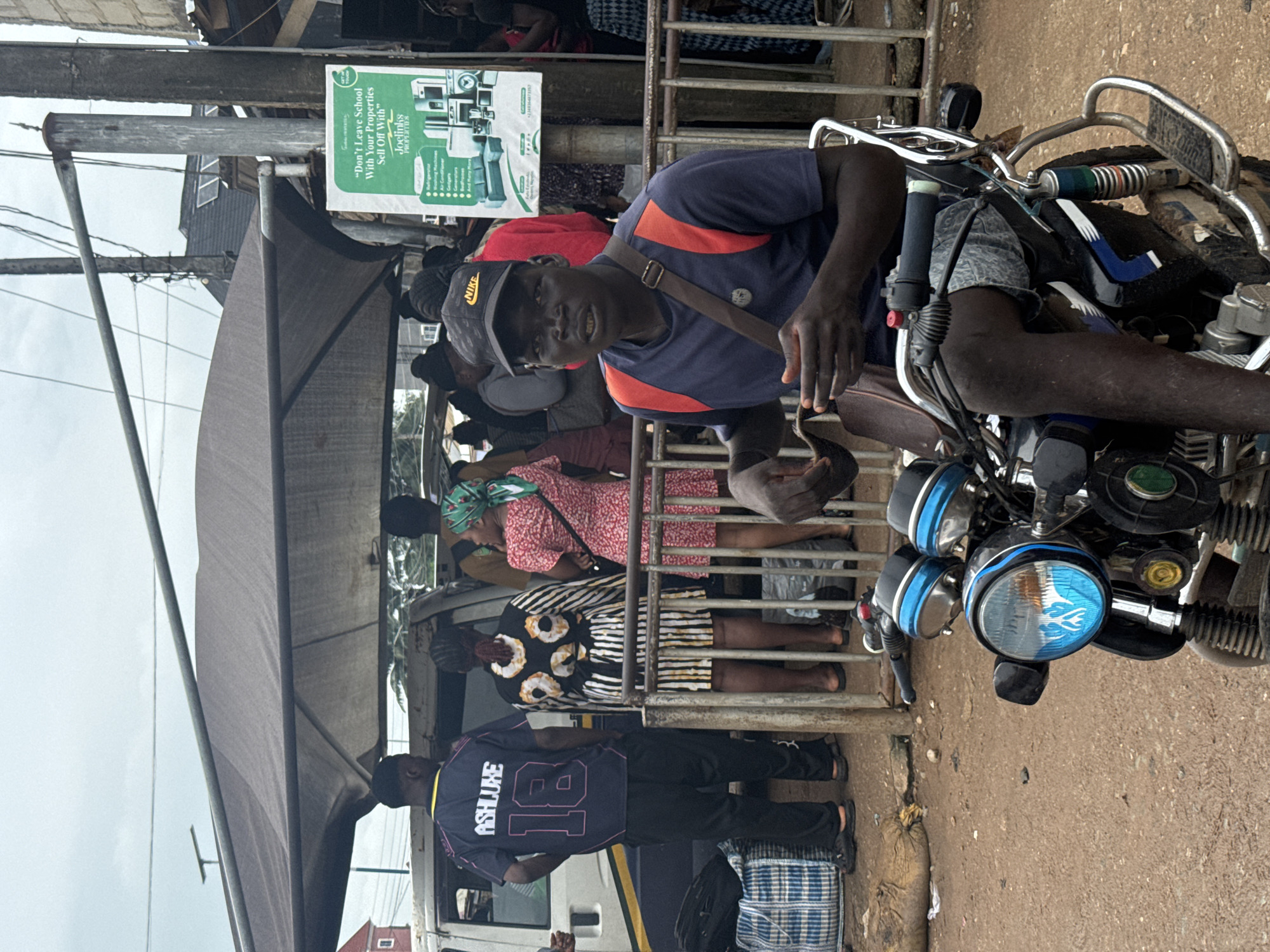 This image captures a vibrant outdoor scene in Ihiagwa, Nigeria, under an overcast sky. In the foreground, a man sits on a motorcycle, engaging with the camera with a slight smile. He is dressed in a navy blue shirt with orange shoulder accents, a black cap bearing the "NIKE" logo, and a shoulder bag. His right hand is raised, making a gesture. The motorcycle has distinctive blue headlights and chrome handlebars.
Behind the man, a metal railing delineates a bustling area where several people are gathered. A white bus or minivan is partially visible, suggesting a transportation hub. Among the crowd, one person is seen from behind, wearing a black jersey with "ASHLUXE" and the number "18" prominently displayed. Another individual wears a patterned dress, standing near the railing.
Above the activity, a large green sign is mounted, partially covered by a dark canopy. Visible text on the sign includes the slogan: "Don't Leave School With Your Properties Sell Off With" followed by "Joelinks PROPERTIES". Below this, a list of various household and electronic items is presented, such as "Refrigerators", "Washing Machines", "Air Conditioners", "Cookers", "Gadgets", "Bed/Foams", and "And Many More". The ground beneath the people and vehicles is a mix of dirt and possibly worn pavement. The setting appears to be a busy, everyday urban environment.