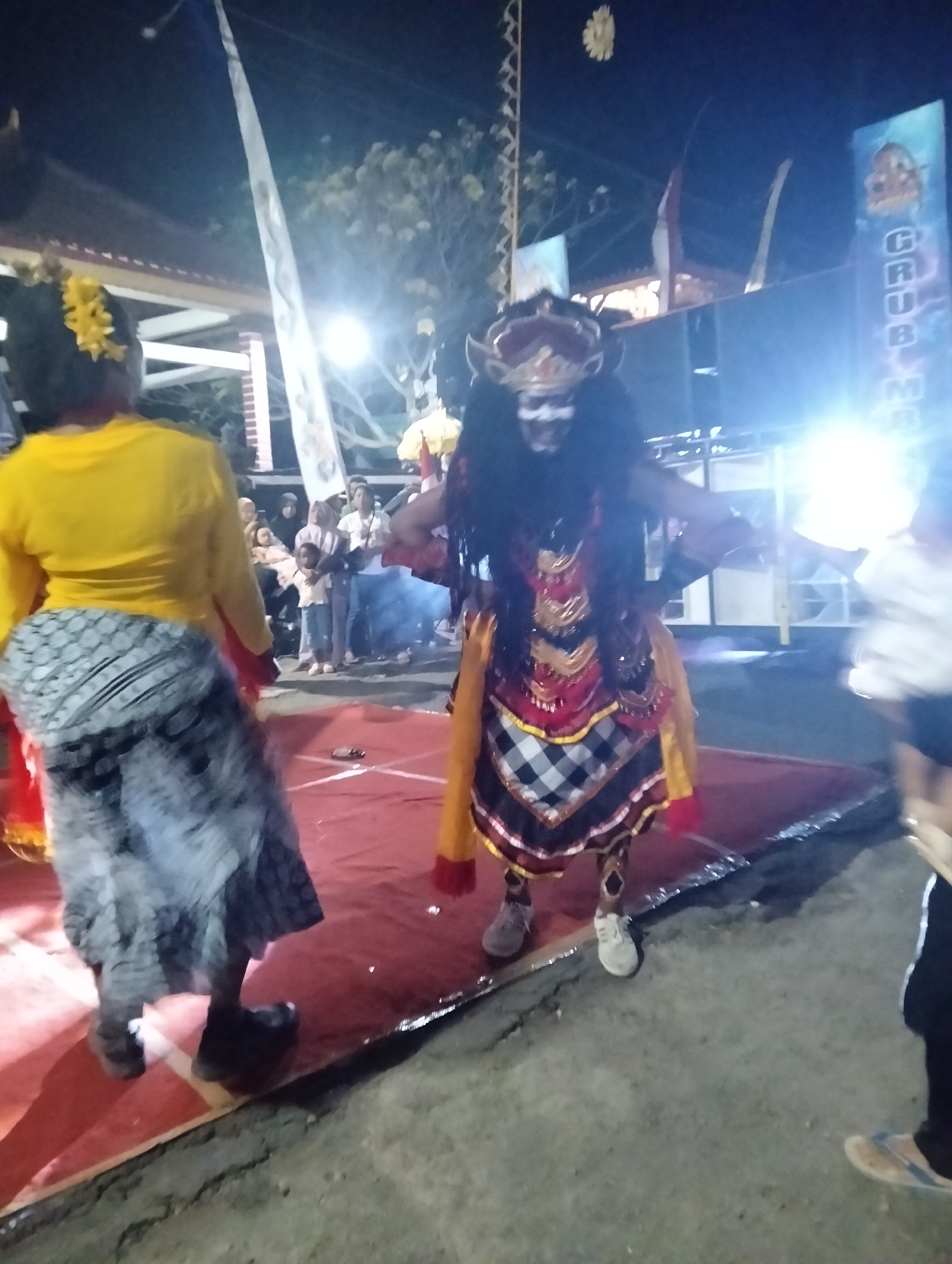 In Sumbersewu, Indonesia, at night, a performance is taking place. A person in elaborate traditional attire, featuring a headdress, dark wig, and a layered costume of red, gold, and black and white checkered fabric, stands center stage. Their face is painted white with dark accents. To their left, another individual in a yellow top and a patterned grey skirt is positioned. In the background, a crowd of spectators, including children, are visible, some standing and others sitting. Various flags and banners are displayed, and bright lights illuminate the area, casting long shadows. The stage itself is a red mat.