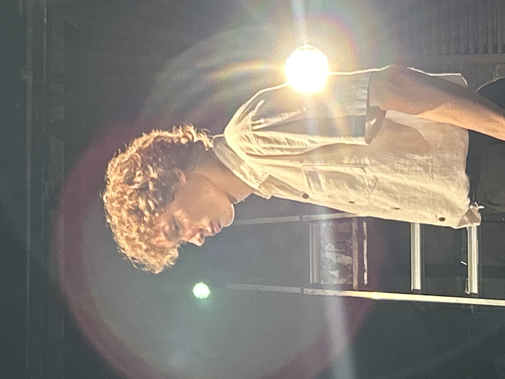 A young male with curly brown hair, wearing a light-colored short-sleeved button-up shirt and dark pants, stands in profile facing left. He is illuminated by bright stage lights, one of which is directly to his right, creating lens flare and a rainbow effect. Behind him, a tall ladder is visible, with its rungs illuminated. The background is dark and appears to be an indoor setting, possibly a stage.
