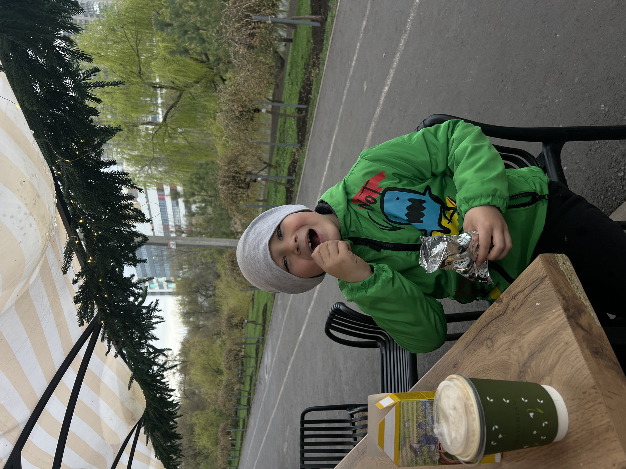A young boy wearing a green jacket and grey beanie is sitting at an outdoor table, appearing to eat or about to eat a wrapped snack. He has his mouth open and is smiling. The table also holds a beverage in a green cup and a small carton. A striped awning and evergreen decoration are visible on the left side of the frame. The background shows a park-like setting with trees and a paved path. The location may be Rostov-on-Don, Russia.