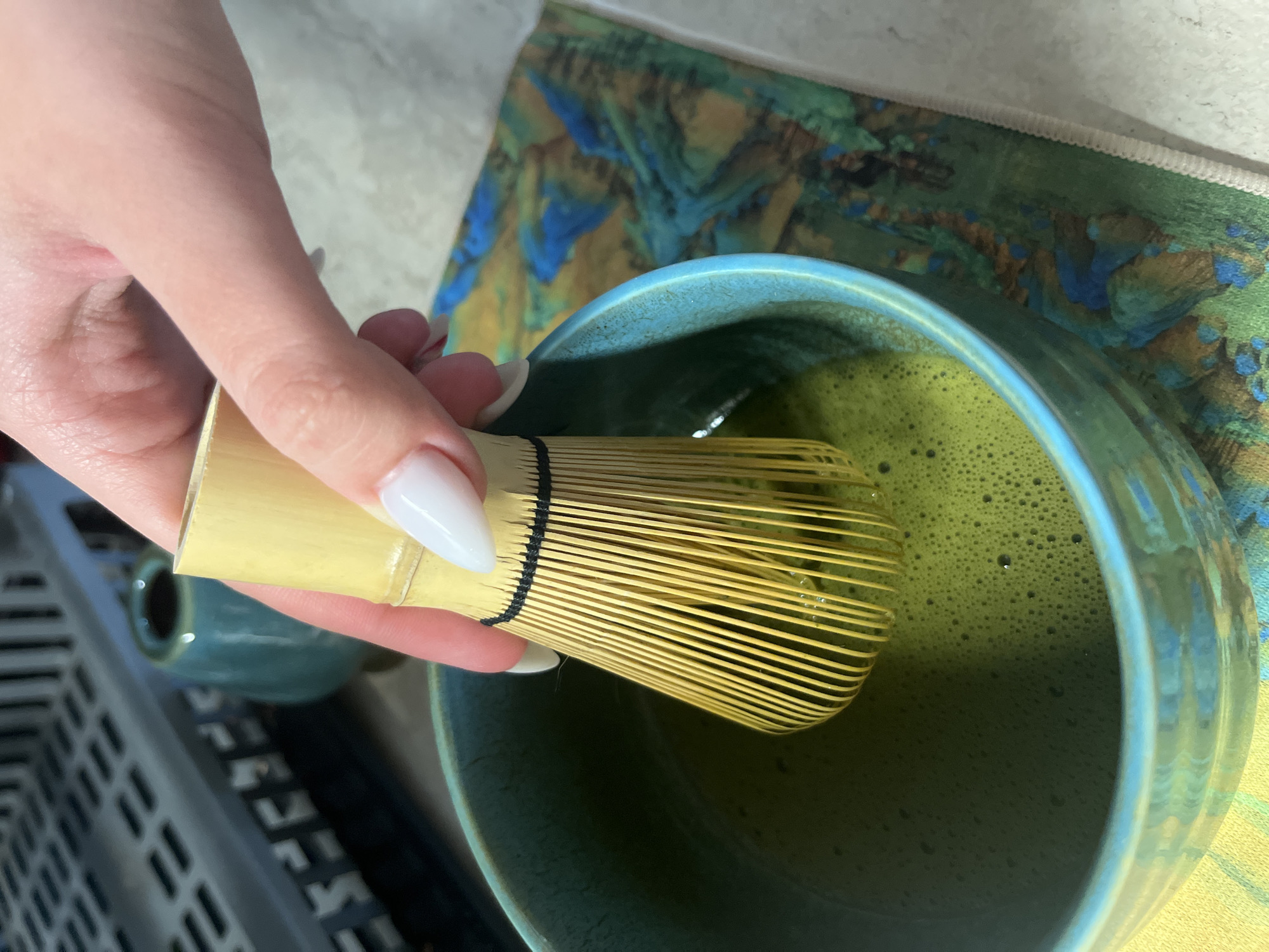 A close-up shot shows a hand holding a bamboo whisk over a blue-green ceramic bowl filled with frothy green matcha tea. The hand has long, pointed white nails. The whisk is positioned in the center of the bowl, partially submerged in the tea. A matching small ceramic cup sits in the blurred background to the left. A patterned cloth in shades of green, blue, and brown is partially visible behind the bowl. The scene is likely indoors and appears to be a preparation of matcha tea. The location, Sofia, Bulgaria, cannot be determined from the visual information provided.