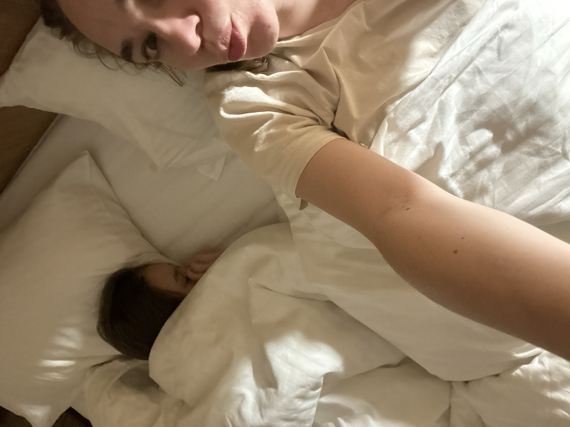 A woman is lying on a bed in a room with a child next to her. The woman is at the top of the image, looking down at the camera with her face visible. Her body is partially covered by a white duvet. The child is in the lower left corner of the image, with their head covered by the white duvet. The background is the bed and white bedding. I cannot discern any information to connect this image with Yekaterinburg, Russia.
