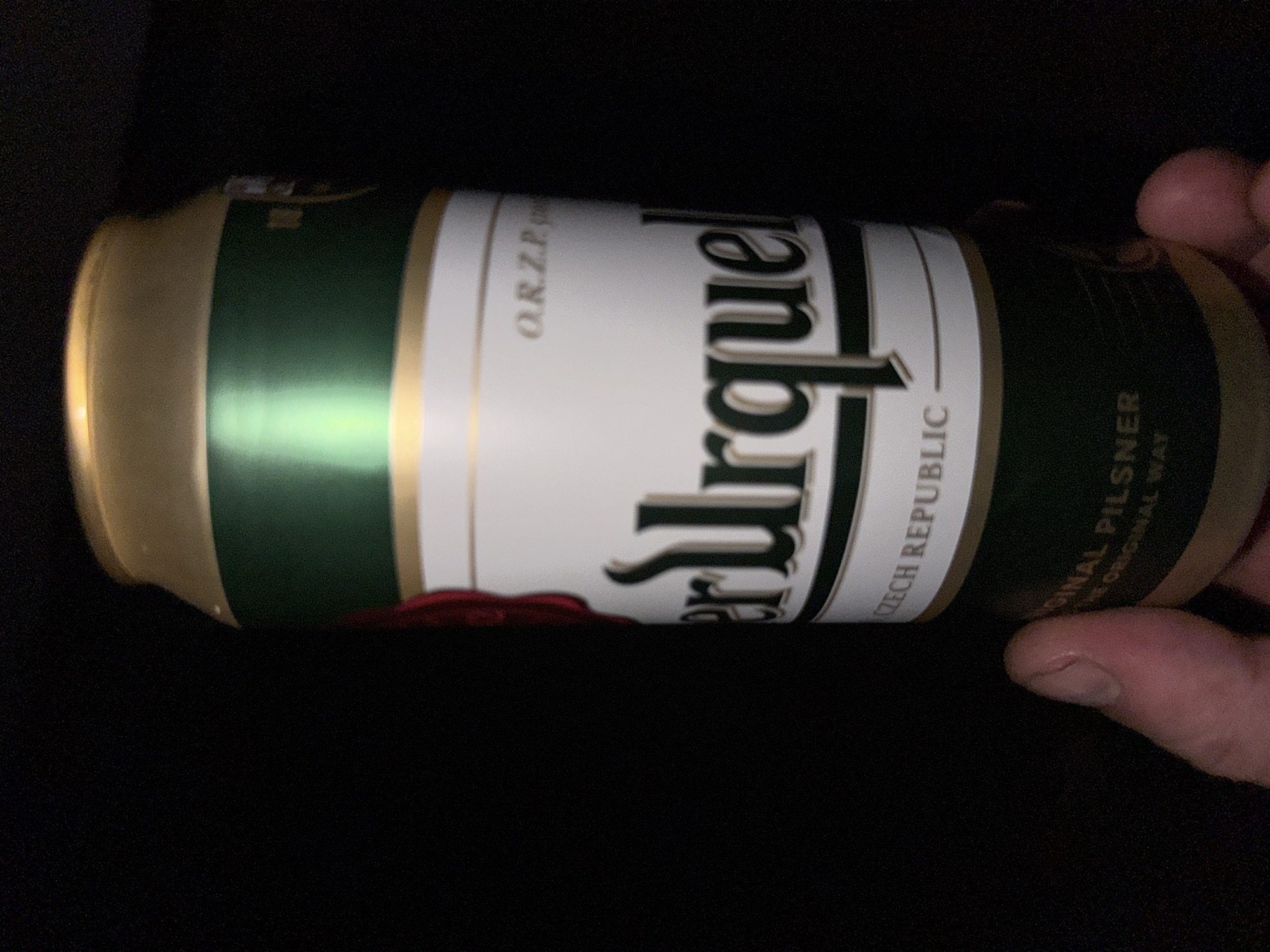 A close-up, eye-level shot shows a person's thumb and forefinger holding a can of Pilsner Urquell beer. The can has a white label with "Pilsner Urquell" in large, dark green lettering. The top of the can is gold and green. The background is dark and out of focus. The scene appears to be indoors.
