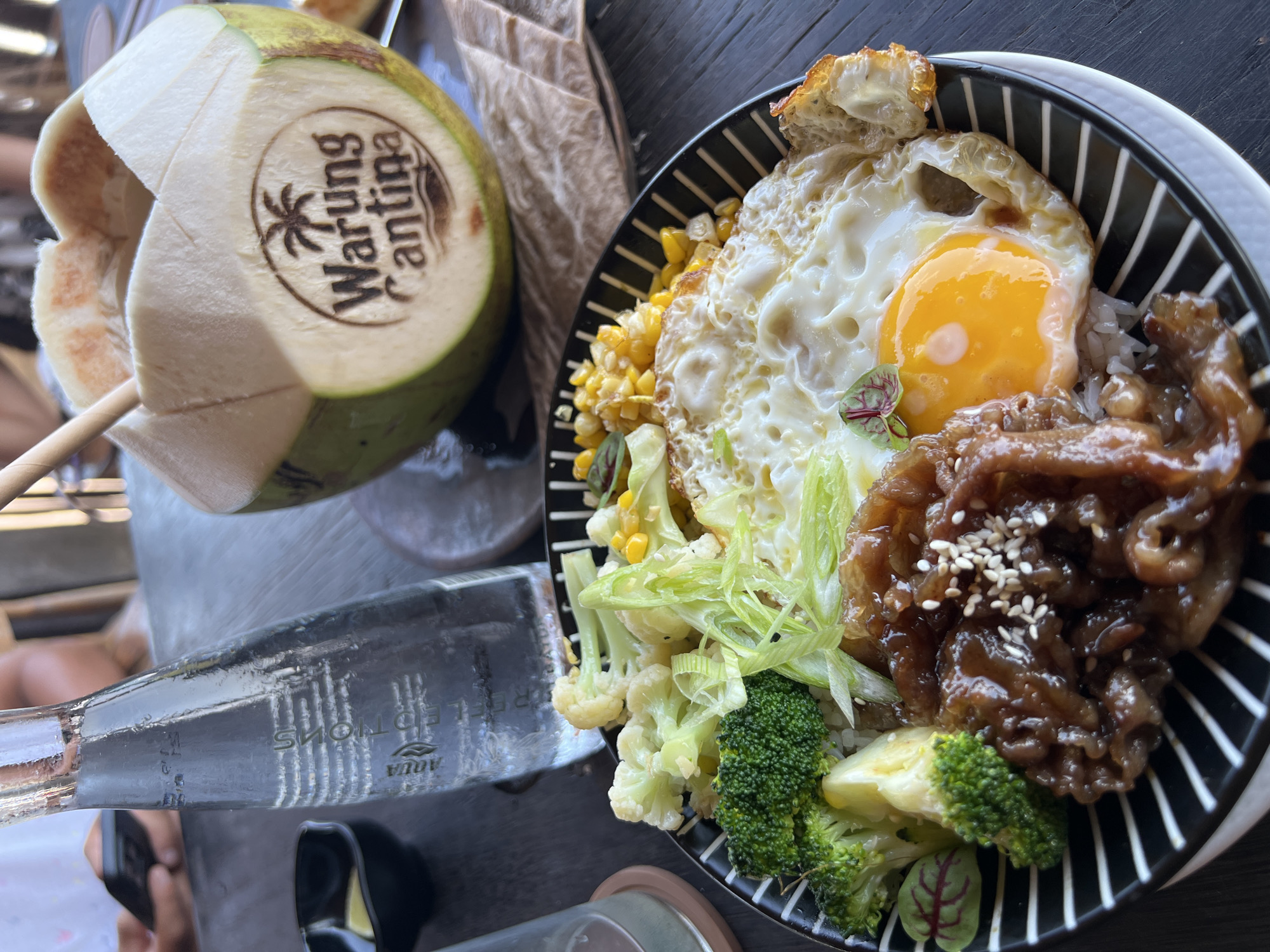 In North Kuta, Indonesia, on a dark wooden table, a meal is served. The dish is presented in a black and white striped bowl and features rice topped with a fried egg, thinly sliced beef in a dark sauce sprinkled with sesame seeds, broccoli, cauliflower, and corn. Beside the bowl, a tall glass bottle of Aqua water stands, partially obscured by the bowl. In the background, a young coconut, branded with "Warung Cantina," is visible, its top cut open to reveal a straw. A person's hand, holding a smartphone, is also visible in the background, suggesting a casual dining or leisure setting. The lighting appears natural, indicating an outdoor or well-lit indoor environment.