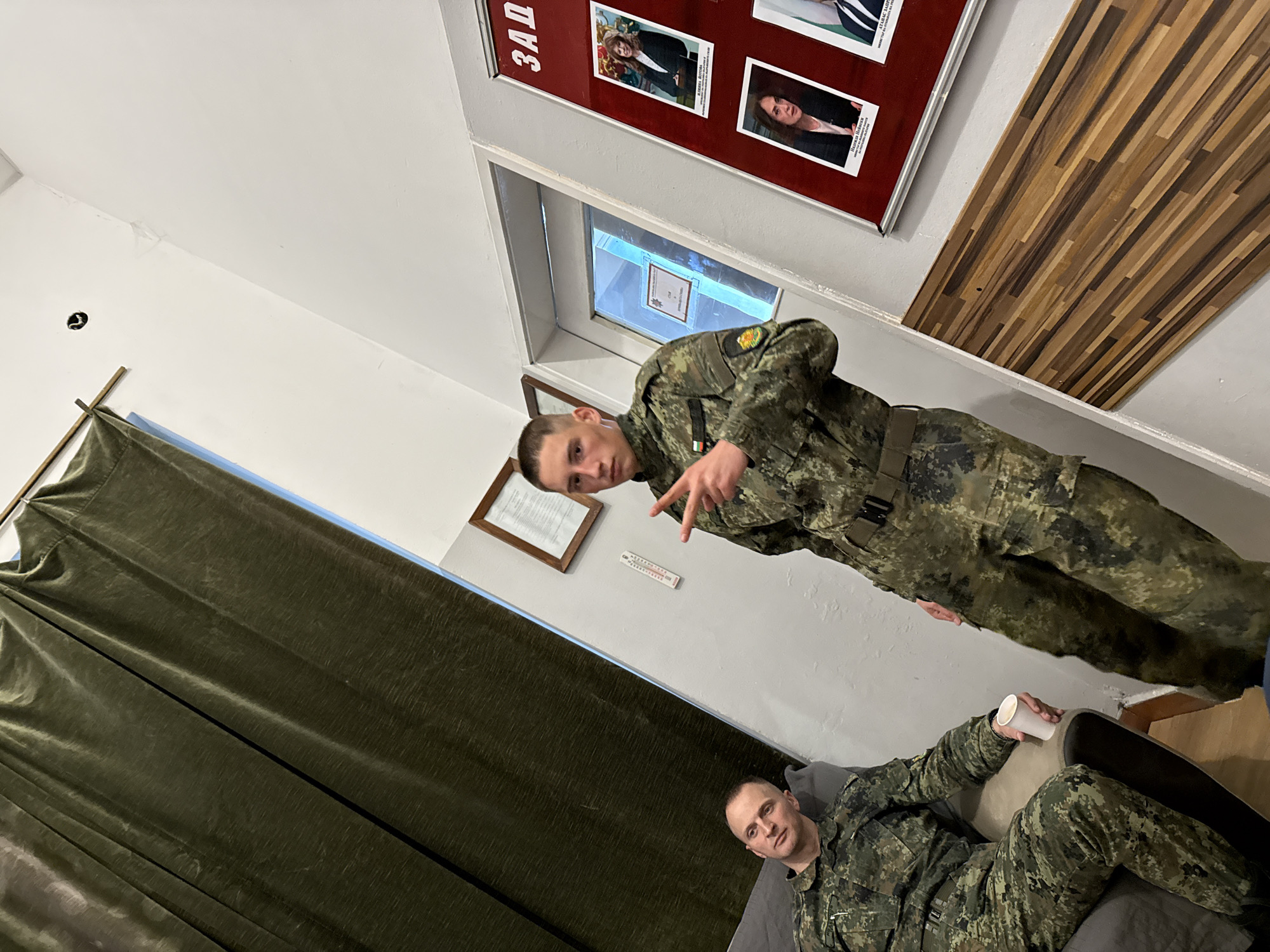Two men in military uniforms are inside a room. One man is standing and gesturing with his hand. The other is seated on a couch holding a cup. The room has green curtains, a picture frame on the wall, a thermometer, and a display board with photos. A sign in Cyrillic is on the display board. The location is identified as Sofia, Bulgaria, based on the possible Cyrillic sign.