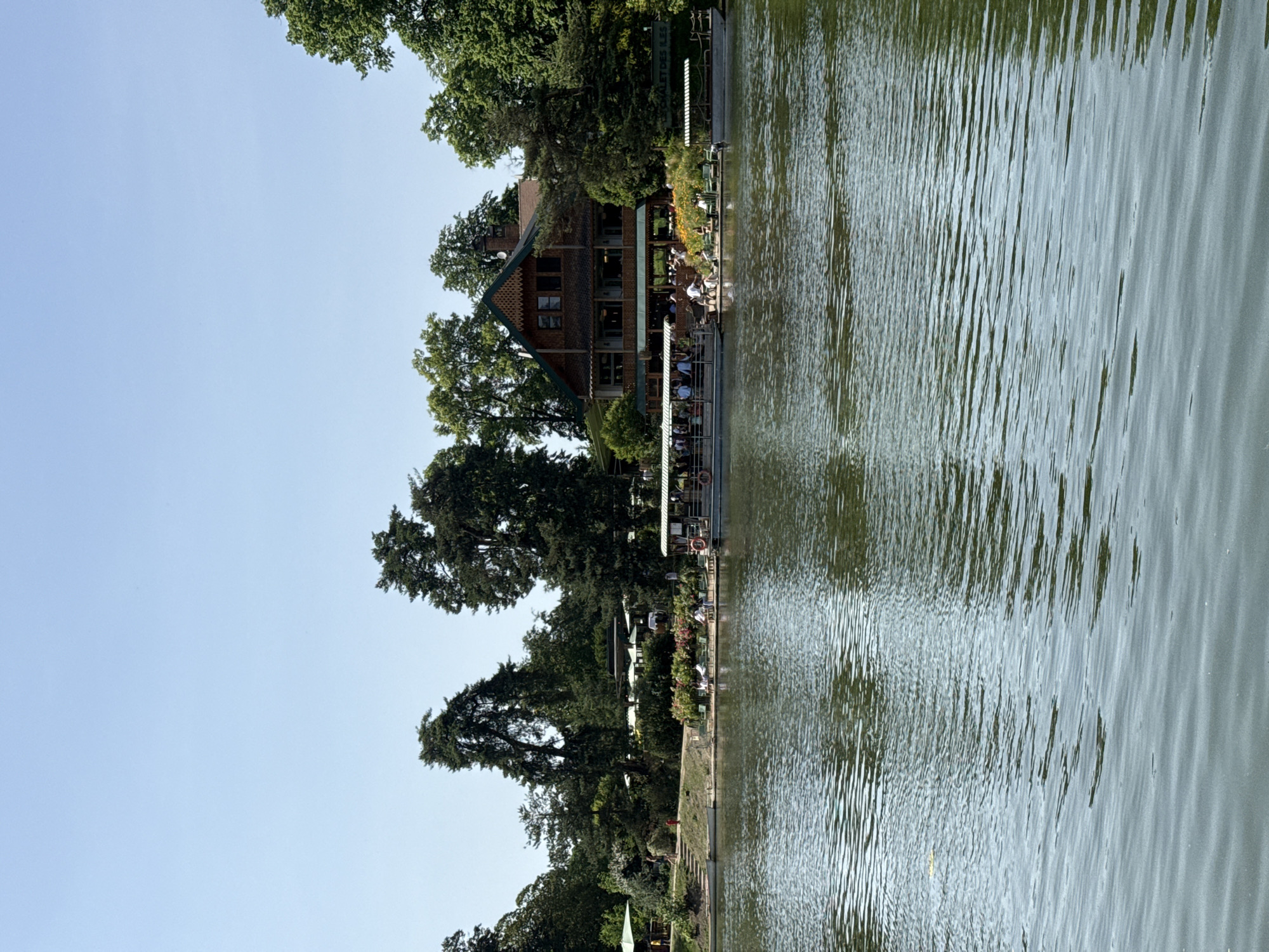 A body of water occupies most of the scene. Along the near bank is a building with a brown exterior, possibly wood, partially obscured by green trees. A lower platform structure is attached to the building, with people sitting and standing near the water. Further along the bank are additional trees, vegetation, and what appear to be recreational areas. The sky above is a pale blue. This is the Chalet des Iles in Paris.