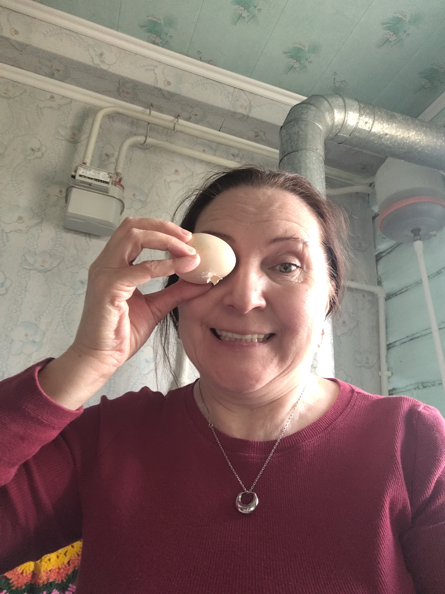 A middle-aged woman is captured indoors, smiling broadly and playfully holding a light brown, partially cracked egg over her right eye. Her dark hair frames a cheerful face, and her visible left eye sparkles with humor. She is wearing a long-sleeved, ribbed maroon top and a silver chain necklace adorned with a unique, oval-shaped silver pendant. A vibrant, multi-colored pattern, possibly knitted or crocheted, is partially visible on her garment at the bottom left.
The setting appears to be a domestic interior in Susady-Ebalak, Russia. The walls are covered with light-toned wallpaper featuring a subtle, light blue floral pattern. The ceiling is lighter, with faint green floral designs, and a white molding separates it from the walls. Utility infrastructure is prominent in the upper background: white pipes run along the wall leading to a white utility meter, which displays small, unreadable text on a label. A large, corrugated metal duct curves from the ceiling, running horizontally before bending downwards. To the far right, a dark grey, circular fixture with a reddish-orange rim and a white pole extends from the ceiling, likely part of a lighting or ventilation system. The scene conveys a lighthearted and candid moment.