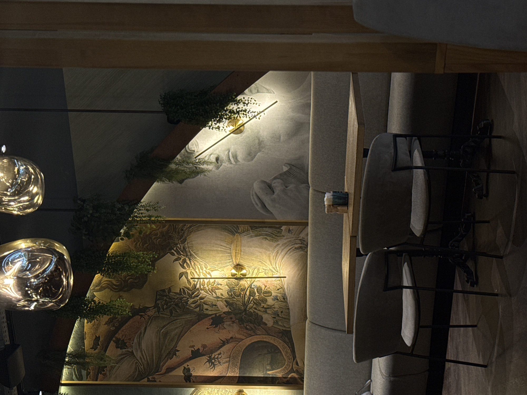 The image depicts the dimly lit interior of a restaurant or cafe in Perm, Russia, characterized by atmospheric lighting and classical-inspired artwork. The scene is presented as if viewed from an elevated angle, looking down into the space.
In the mid-ground, a prominent wall on the left features a large mural-style painting in a classical or Renaissance aesthetic. This artwork portrays a figure, possibly female, surrounded by lush foliage, intricate flowers, and botanical motifs, illuminated by a linear light fixture positioned above it. Green, artificial or preserved foliage is draped along wooden beams and integrated into the wall decor, appearing around the mural. Two large, irregularly shaped, reflective pendant lamps hang from the ceiling on the far left, adding to the sophisticated lighting scheme.
To the right, upholstered booth seating with gray cushions runs along another wall, complemented by several gray upholstered chairs with black metal frames, positioned around narrow wooden tables. Further along this wall, a second mural depicts the stylized face of a classical figure, illuminated by a similar linear light fixture. More green foliage is visible near this artwork, continuing the natural decorative theme.
The overall lighting is warm and artificial, suggesting an evening or nighttime setting, creating a dramatic and intimate ambiance. No people are visible, indicating the establishment is either empty or awaiting patrons. There is no visible text in the image.