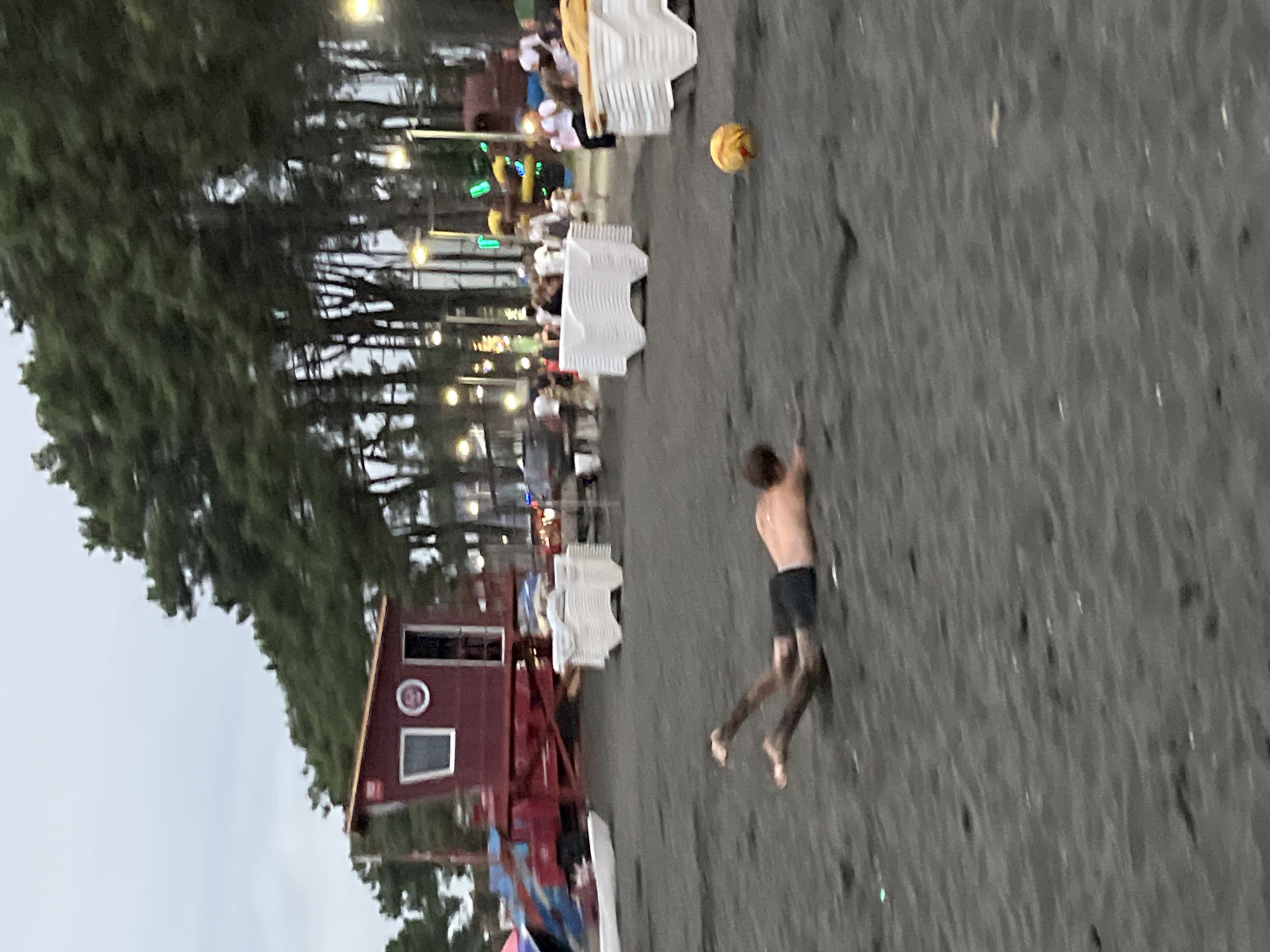 The image captures a scene on the beach in Ureki, Georgia, known for its black magnetic sand. In the foreground, a person is lying prone on the sand, appearing to be in motion, with their legs extended and torso parallel to the ground, suggesting they might be sliding or falling. They are wearing dark shorts. The sand itself is a dark, granular material. In the mid-ground, several white plastic chairs are stacked neatly, and a yellow ball is visible on the sand. Behind the chairs, a red lifeguard tower with a window and a circular emblem is situated. A vehicle with a red and blue structure is parked behind the tower. The background features a line of tall, slender trees with dense green foliage, and beyond them, streetlights and more trees are visible, indicating an area with some development. The sky is overcast and light gray. The overall impression is of a beach during what appears to be the late afternoon or early evening, with some recreational activity and infrastructure present.