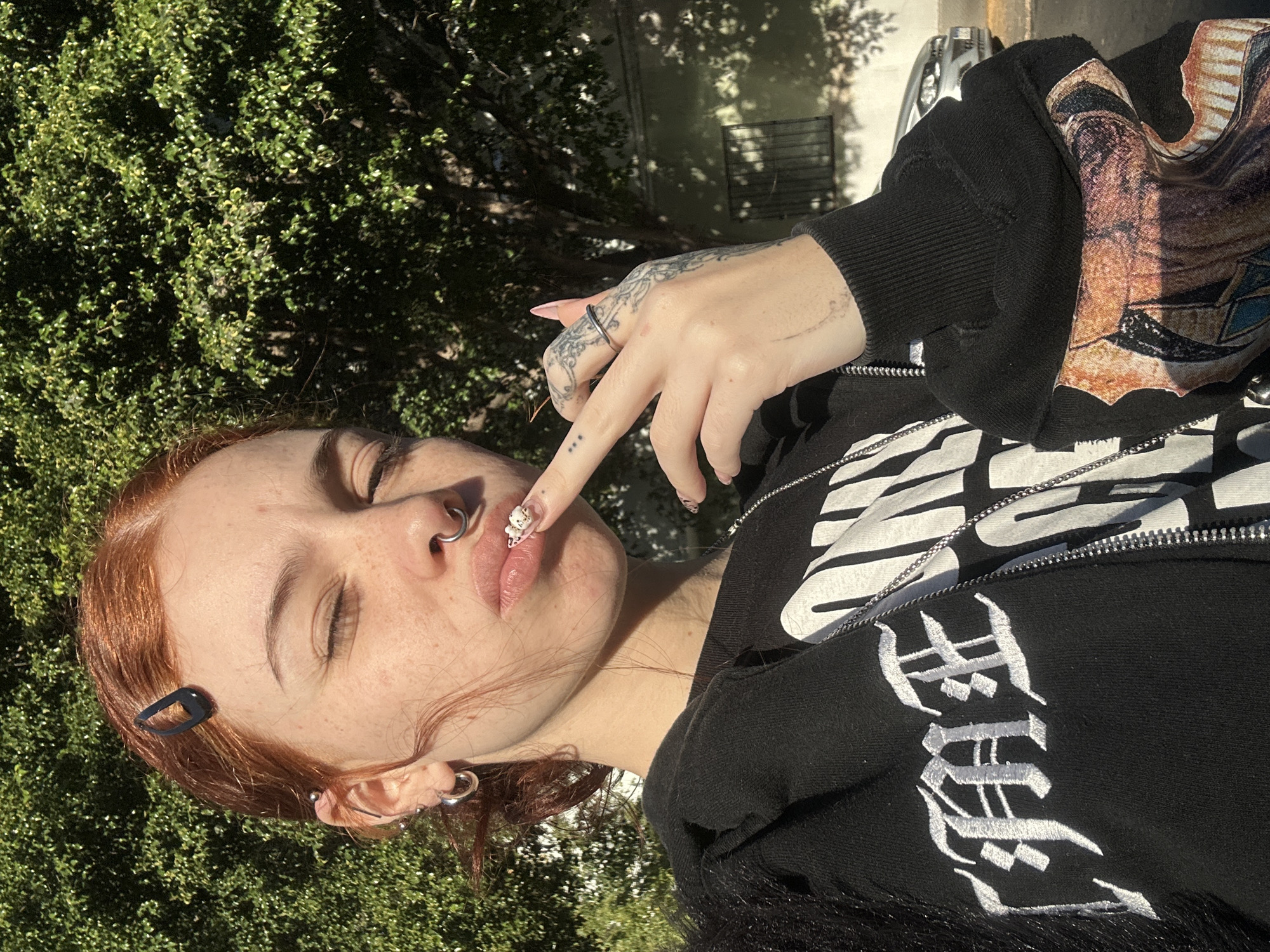 A horizontally oriented outdoor shot captures a young person with reddish-brown hair, fair skin, and freckles, against a backdrop of dense green foliage under bright daylight in Buenos Aires, Argentina. The subject's eyes are closed or squinting, possibly due to the sun. They sport multiple piercings, including a septum ring, a nostril ring, and several ear piercings (helix, tragus, and multiple lobe rings). A black hair clip is visible on the right side of their head.
The person is wearing a black zip-up hoodie featuring white embroidered text in a Gothic font on the sleeve, which appears to spell "Hell". A white graphic print is visible under the zipper, and a colorful, patterned fabric is seen on the far right. Their right hand is raised, with the index finger touching their lips. The hand is adorned with intricate tattoos and a silver ring on the ring finger, while the index finger has three tattooed dots. The long, pink nails on the index finger feature elaborate 3D white charms. The strong sunlight casts distinct shadows, highlighting the vibrant greenery of the outdoor setting, likely a park or garden.