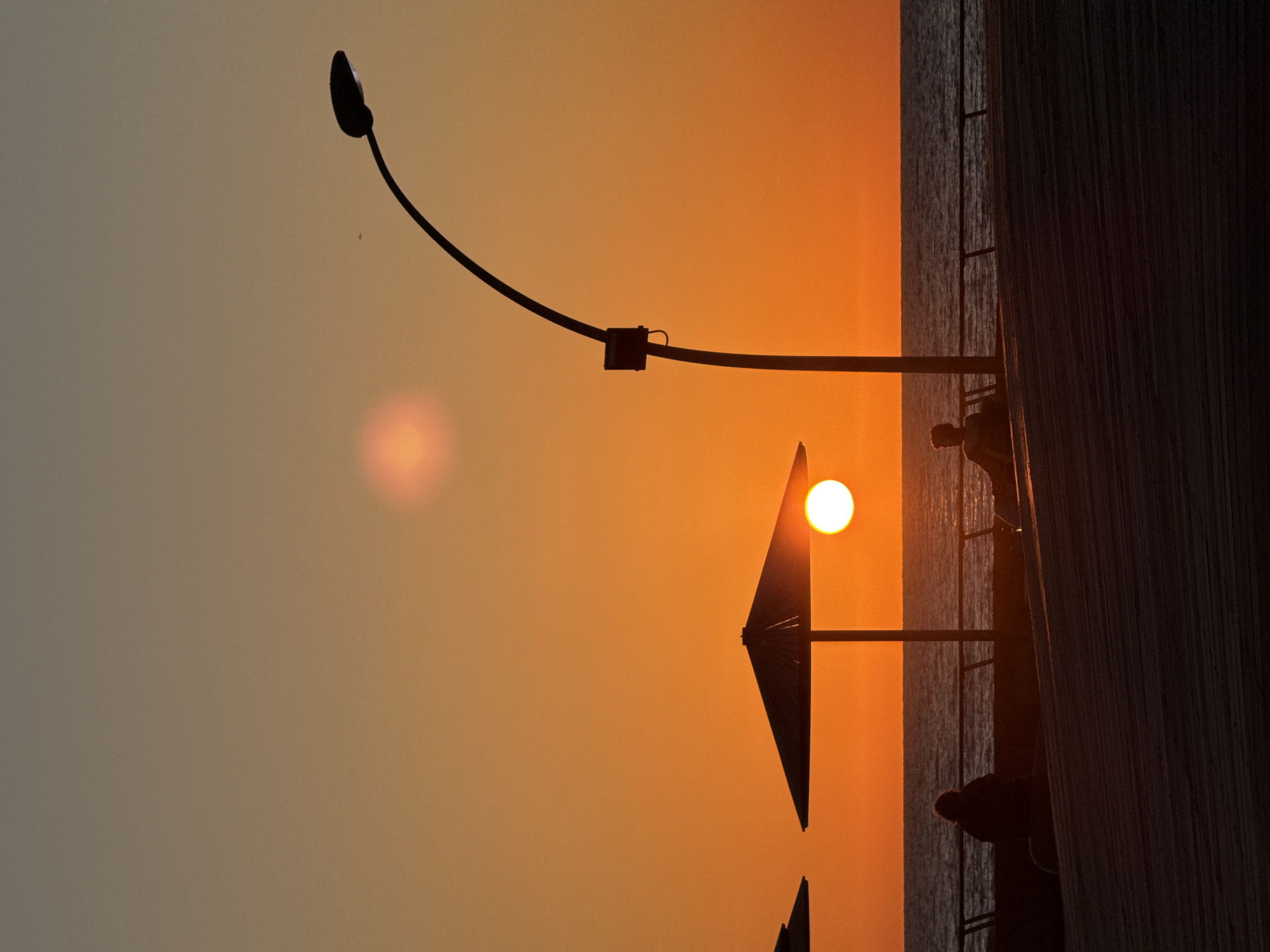 This image captures a serene sunset or sunrise over a body of water, likely the sea, casting foreground elements into stark silhouette. The sky dominates the frame, showcasing a warm gradient from a bright, vibrant orange-yellow near the horizon to a softer, hazy gray-brown further away. A brilliant, circular, white-yellow sun is prominently featured, positioned towards the lower left of the frame (if oriented horizontally). A fainter, reddish-orange light flare or reflection is visible above and to the left of the sun.
Several objects and figures are silhouetted against the bright sky. A long, gracefully curved streetlight extends from the top right, with its lamp head visible at the frame's edge. Below the streetlight and to the sun's left, the distinct triangular shape of a large beach umbrella or parasol stands out, with hints of a second one below it.
On the right side of the image, a calm expanse of water, likely the Mediterranean Sea given the Tel-Aviv context, exhibits subtle ripples on its surface. Adjacent to the water, a dark, textured vertical structure, possibly a wooden wall or pillar of a promenade or pier, occupies the far right. Between this structure and the water, faint lines suggest a ladder or railing. Several dark human silhouettes are visible near the water and the structure, appearing to observe the scenic view. One figure is clearly distinguishable from the chest up, and another's head is visible closer to the bottom edge.
The atmosphere conveyed is peaceful and contemplative, capturing a tranquil moment at dusk or dawn. The image is currently displayed rotated 90 degrees clockwise from its likely original landscape orientation. There is no visible text.