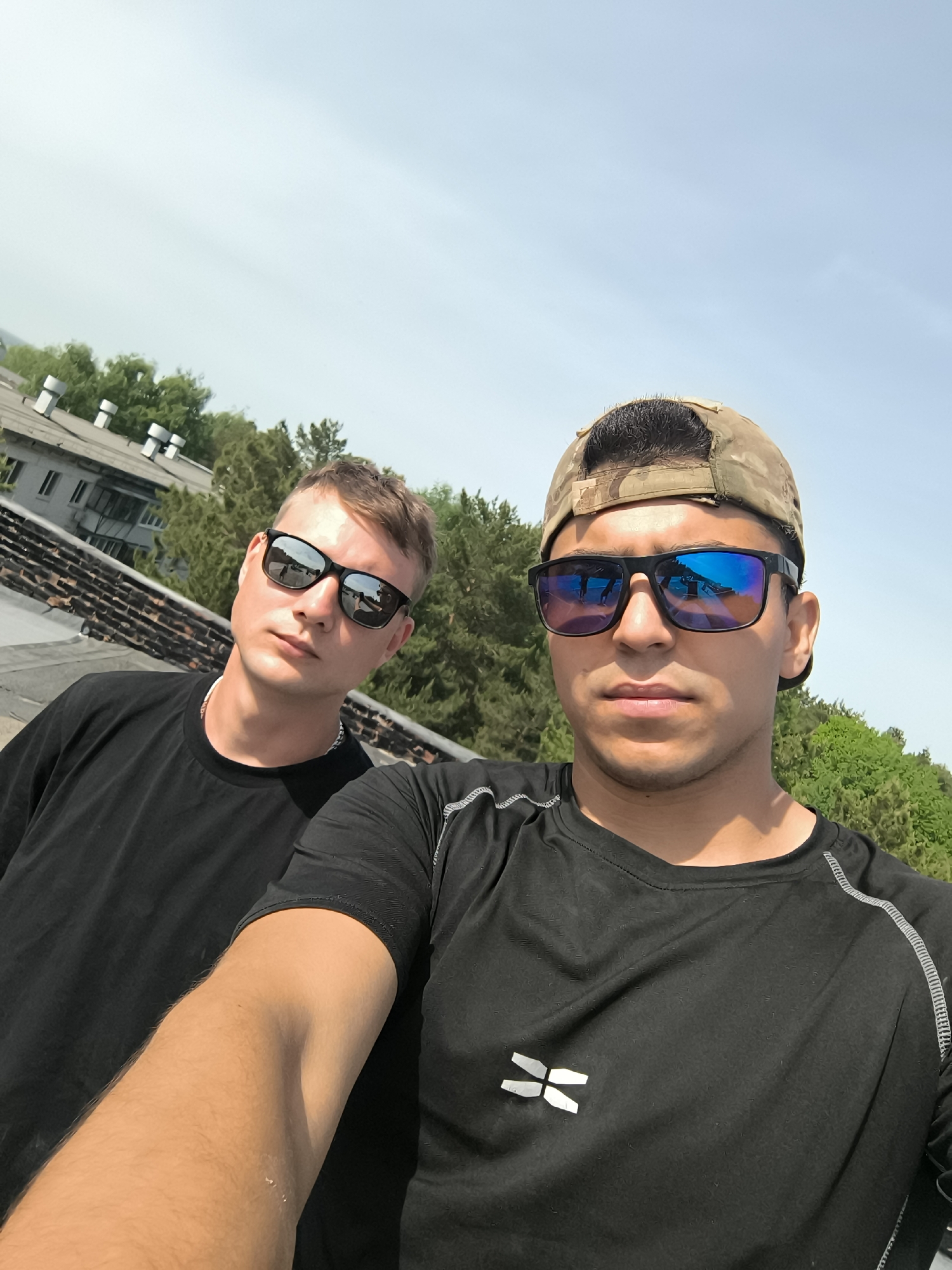 Two males stand side-by-side, taking a selfie outdoors. The individual in the foreground wears a black, long-sleeved shirt with a white logo, a camouflage baseball cap, and dark sunglasses with blue-tinted lenses. The second male is slightly behind and to the left, wearing a black t-shirt and dark sunglasses. The background includes a brick-covered rooftop, a building with chimney-like structures, and green trees against a light blue sky.