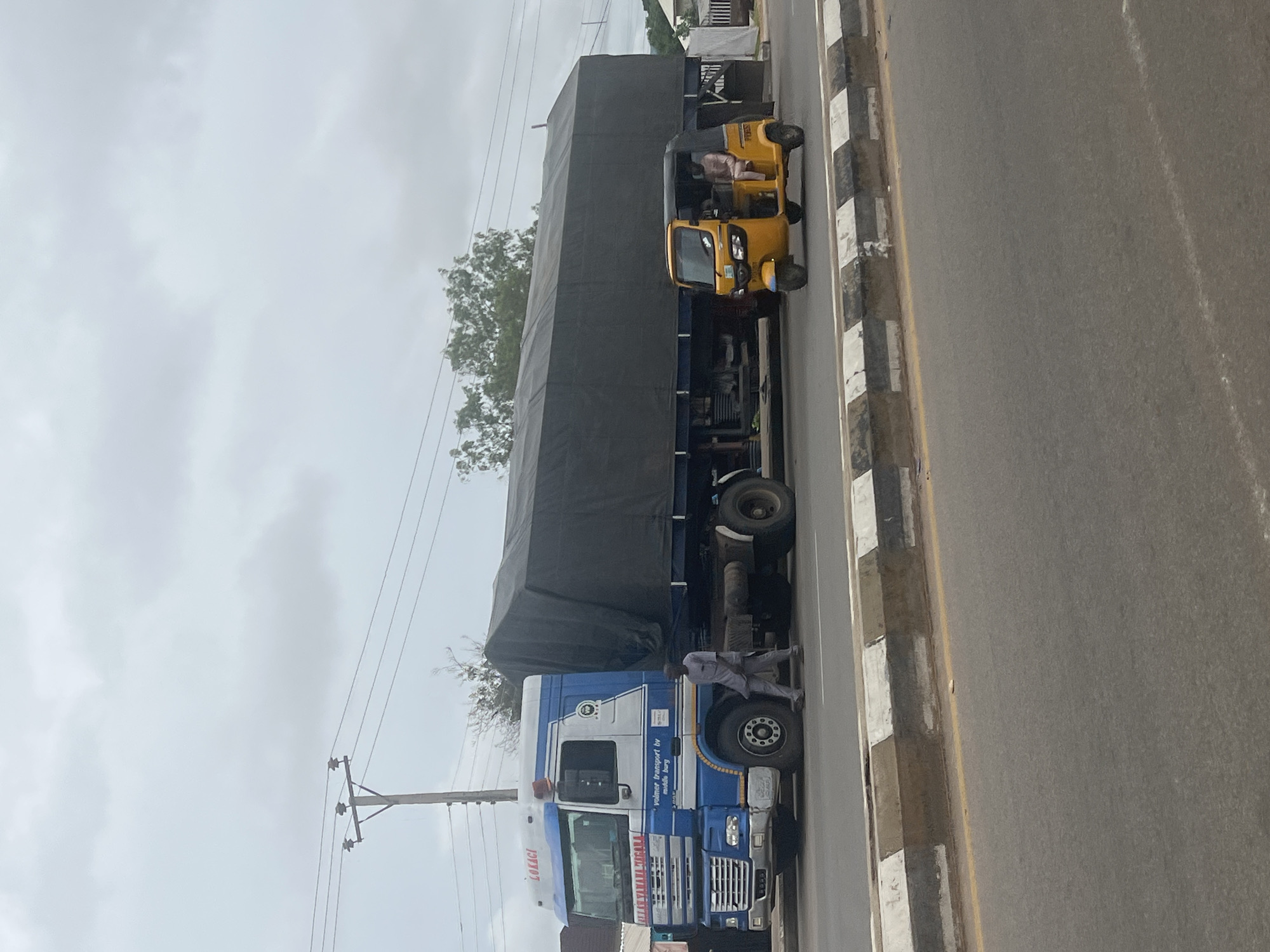 The image depicts a street scene in Jalingo, Nigeria, under an overcast sky. In the foreground, a paved road is visible with a concrete curb marked with black and white stripes. A yellow auto-rickshaw, also known as a Keke Napep, is in motion on the road, carrying two occupants. To the left of the auto-rickshaw, a large blue and white truck with a black tarp-covered cargo trailer is parked or moving slowly. A man dressed in a grey outfit is standing near the truck. A utility pole with wires stretches across the upper portion of the image. In the background, trees and a few buildings are visible, suggesting an urban or semi-urban environment.
