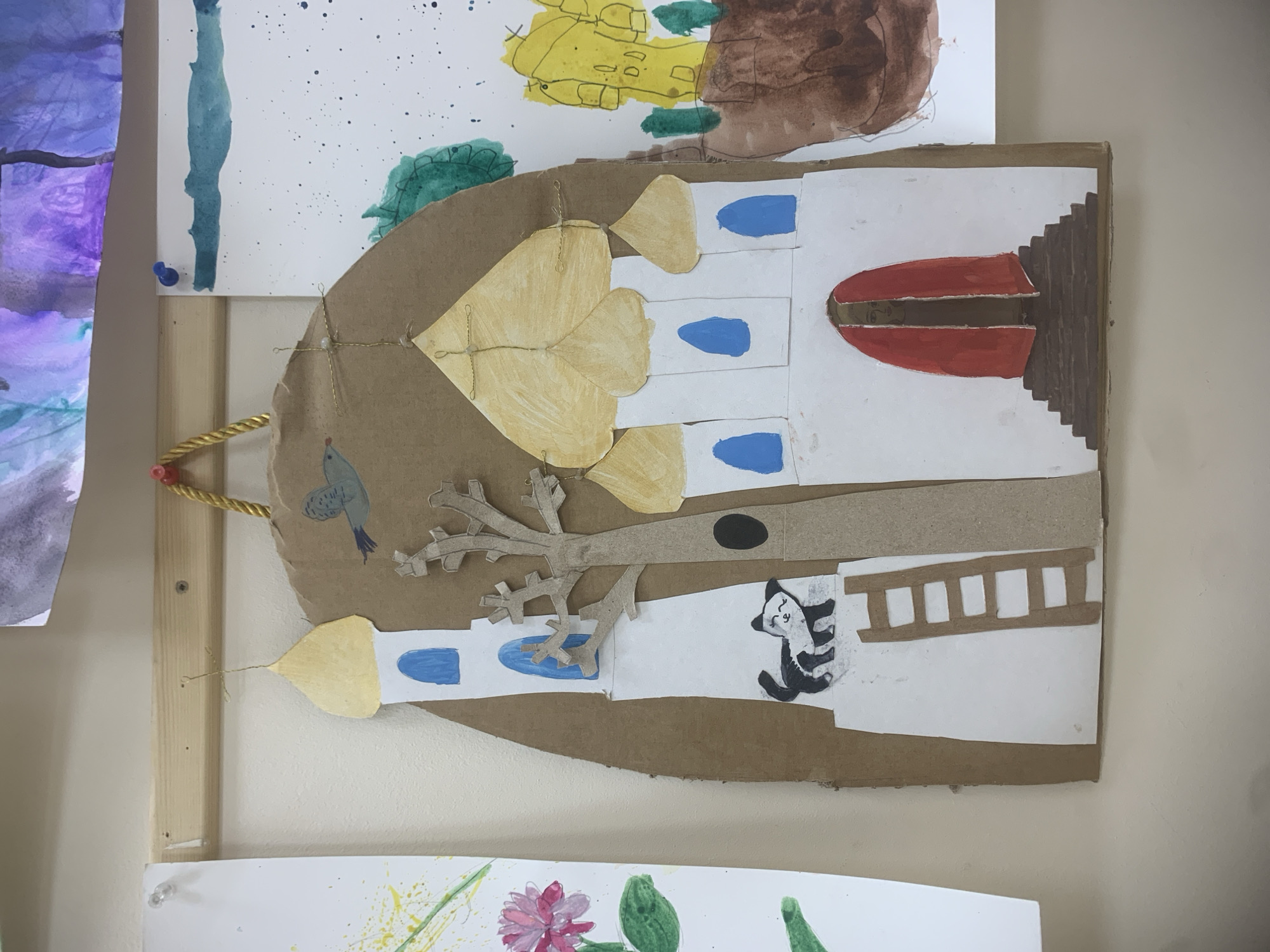 A child's artwork, seemingly a construction of a building or structure resembling a Russian Orthodox church with onion domes, is displayed on a wall. The artwork utilizes cardboard as the base and incorporates paper cutouts and drawings. The overall shape is bell-like. The building features white elements with blue accents and a red, eye-like opening. A small, sketched cat-like figure is visible near what seems to be a ladder-like structure. There's a tree branch made of cardboard attached to the facade. It is unclear if this artwork depicts a real location in Izhevsk, Russia.