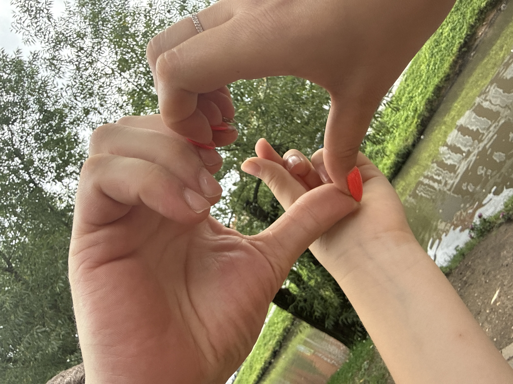 Two people's hands are forming a heart shape outdoors, with a body of water and green foliage in the background, likely in a park or natural setting. The hands are close to each other, with one person's thumb touching another's index finger to complete the heart. There is a thin red string visible. One person is wearing a ring on their ring finger. The context of Dmitrov, Russia is not discernible from the image itself.