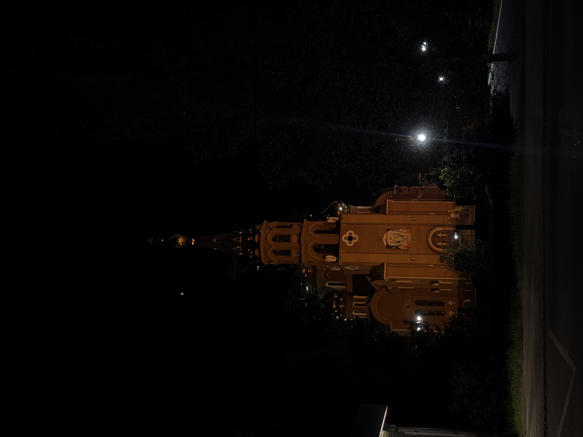 The image shows the Church of the Holy New Martyrs and Confessors of Russia in Chaikovsky, Russia, at night. The church, with its distinctive spire and illuminated facade, stands out against the dark sky and surrounding trees. A street runs along the right side of the image. Streetlights are visible in the background.