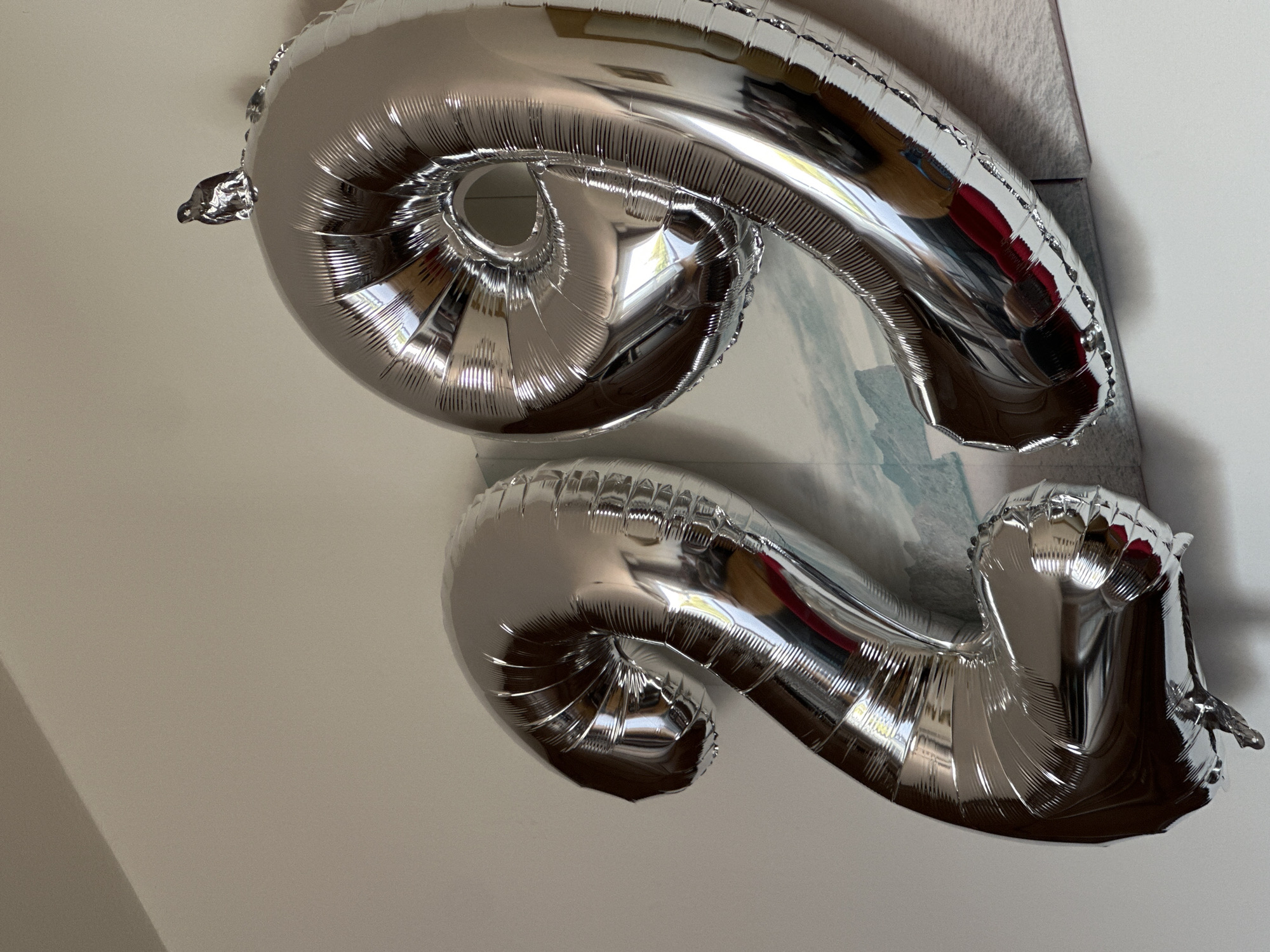 Two large, silver, foil number balloons, shaped like a "2" and a "9", are attached to a wall. The "2" balloon is on the left and the "9" balloon is on the right. Both balloons are inflated and have a shiny, reflective surface that shows distorted reflections of the surrounding environment. The "9" balloon has a small, dark object, possibly a piece of tape or a small decoration, adhered to its top curve. The wall behind the balloons is painted a light, neutral color and appears to have a patterned poster or mural hanging behind the balloons, depicting a mountainous landscape. The image is a close-up shot, focusing on the balloons and the wall. The scene is indoors, with the lighting suggesting it is daytime. There are no people visible in the image. The location in the city of Grad Rijeka, Croatia cannot be determined from the visual information provided.