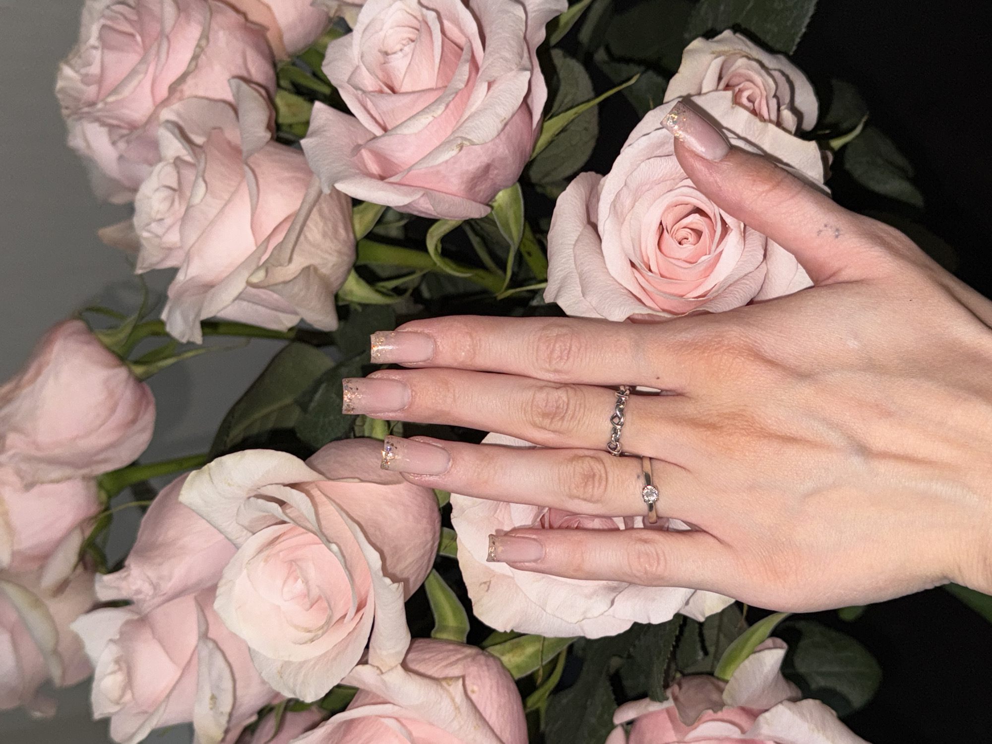 In this close-up, indoor shot, a person's hand, adorned with a light pink, sheer nail polish with glitter accents and two rings, is held in front of a bouquet of light pink roses. One ring is a delicate band with a small diamond, while the other is a silver band with a heart-shaped design. The roses are in various stages of bloom, with soft petals and green stems. The lighting is dim, creating a romantic and intimate atmosphere. The background is dark, further emphasizing the subjects. This image does not provide any information about the city of Perm, Russia.