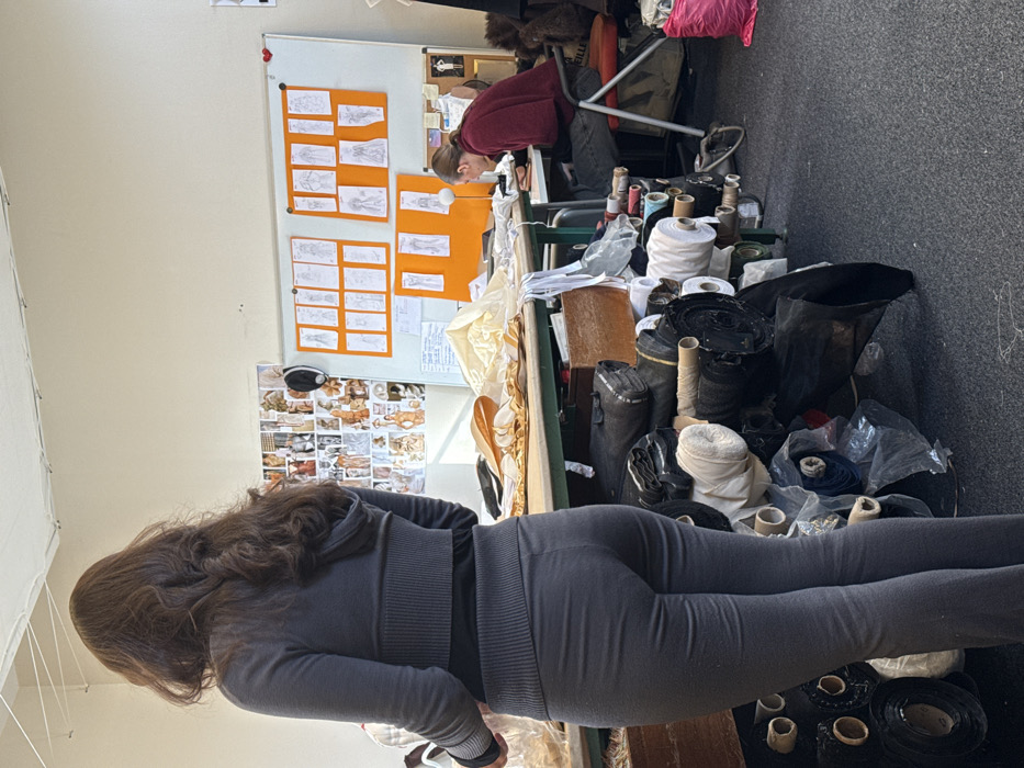 This image depicts an indoor workshop or design studio located in Arcueil, France. The scene features two individuals and numerous items related to garment design or production.
In the foreground, slightly to the left of the center, a female subject stands with her back predominantly to the viewer, facing towards the right side of the frame. She has long, dark brown, wavy hair. She wears a dark grey long-sleeved top with a ribbed waistband and matching dark grey fitted trousers. Her right arm is slightly lifted, positioned over a work surface.
To the right, in the background, a second individual, appearing male, is seated at a table, facing towards the left. He has short dark hair pulled back. He wears a dark red long-sleeved top and dark trousers. He is leaning forward, focused on items on the table in front of him. He is seated on a chair with a red back and an orange seat, upon which the word "ARCUEIL" is partially visible.
The room has light-colored, possibly off-white, walls and a dark grey carpeted floor. Across the ceiling, white electrical wires are visibly strung. Along the back wall, to the left of the seated individual, a large white whiteboard displays several sheets of orange paper, each containing multiple hand-drawn fashion sketches of figures in various poses and attire. To the left of the whiteboard, a collage of smaller images, likely reference photographs or mood board elements, is affixed to the wall.
A large, light-colored work table stretches across the mid-ground. Its surface is covered with folded cream or gold-colored fabric and various white strips of material. Beneath this table and on the floor in the foreground, numerous rolls of fabric are stacked and piled. These rolls vary in color, including black, white, cream, blue, and dark grey, with some wrapped in clear plastic. A wooden box is also visible among the fabric rolls under the table. To the far right, on the floor, a pink plastic bag is present. The space is illuminated by overhead lighting and ambient light from an unseen source.