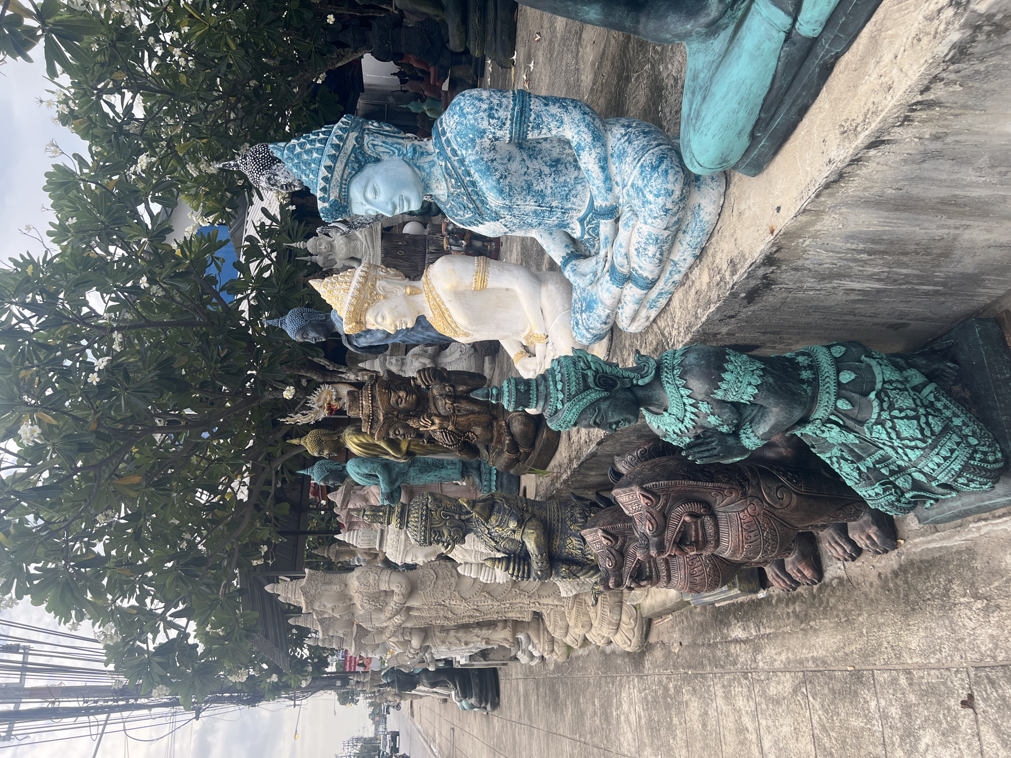This is a full shot of a Buddha statue shop in Ko Samui, Thailand. The shop is located on the side of a road, with a sidewalk and a street visible in the background. There are many Buddha statues of various sizes and colors displayed on the sidewalk and on shelves. The statues are made of different materials, including stone, ceramic, and metal. Some of the statues are very detailed and ornate, while others are simpler. The shop appears to be outdoors, with a roof and some trees providing shade. The sky is overcast, and there is a light breeze.